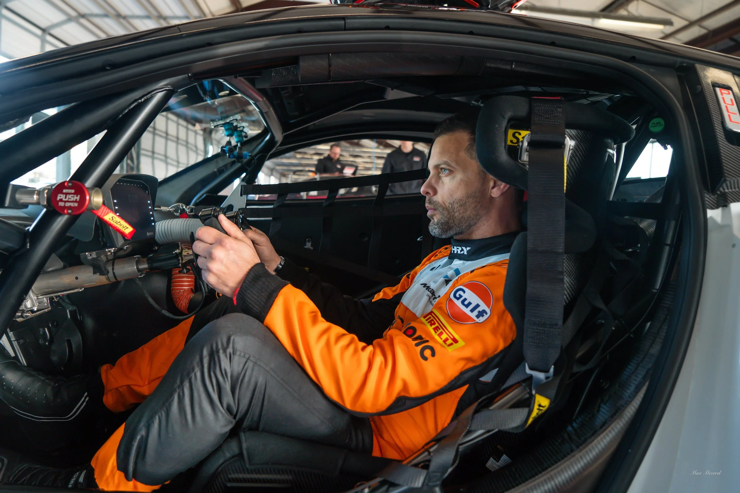 Race car driver sitting inside a race car, wearing an orange racing suit with sponsor logos, focusing on the steering wheel.