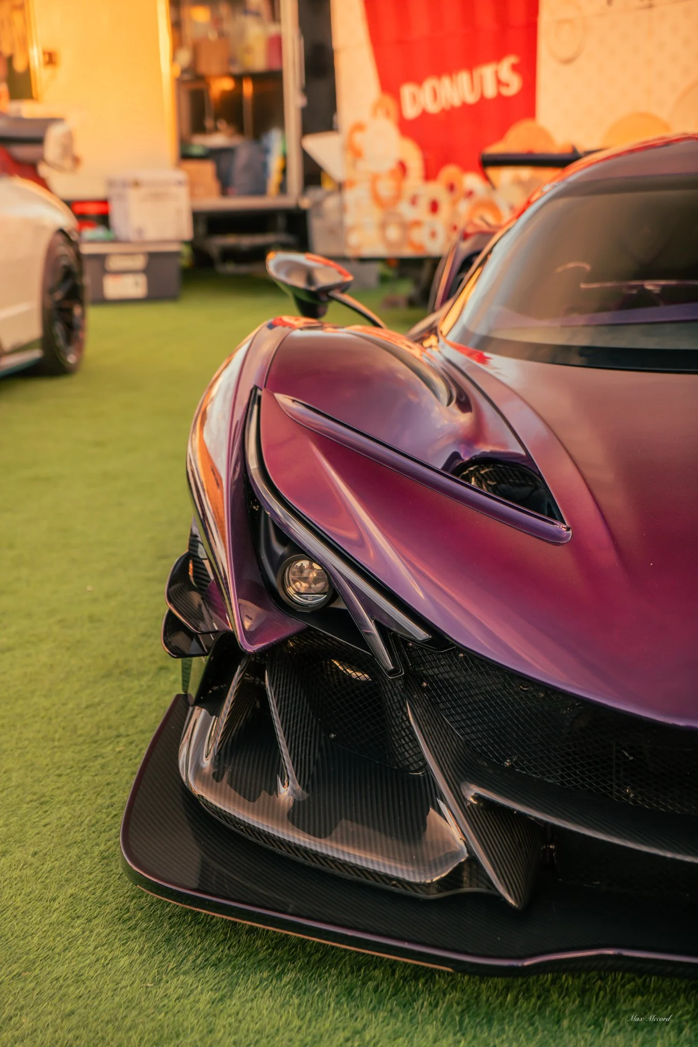 Close-up of the front end of a sleek, purple sports car with aerodynamic features, carbon fiber accents, and a prominent headlight, parked on green artificial grass at an outdoor event.