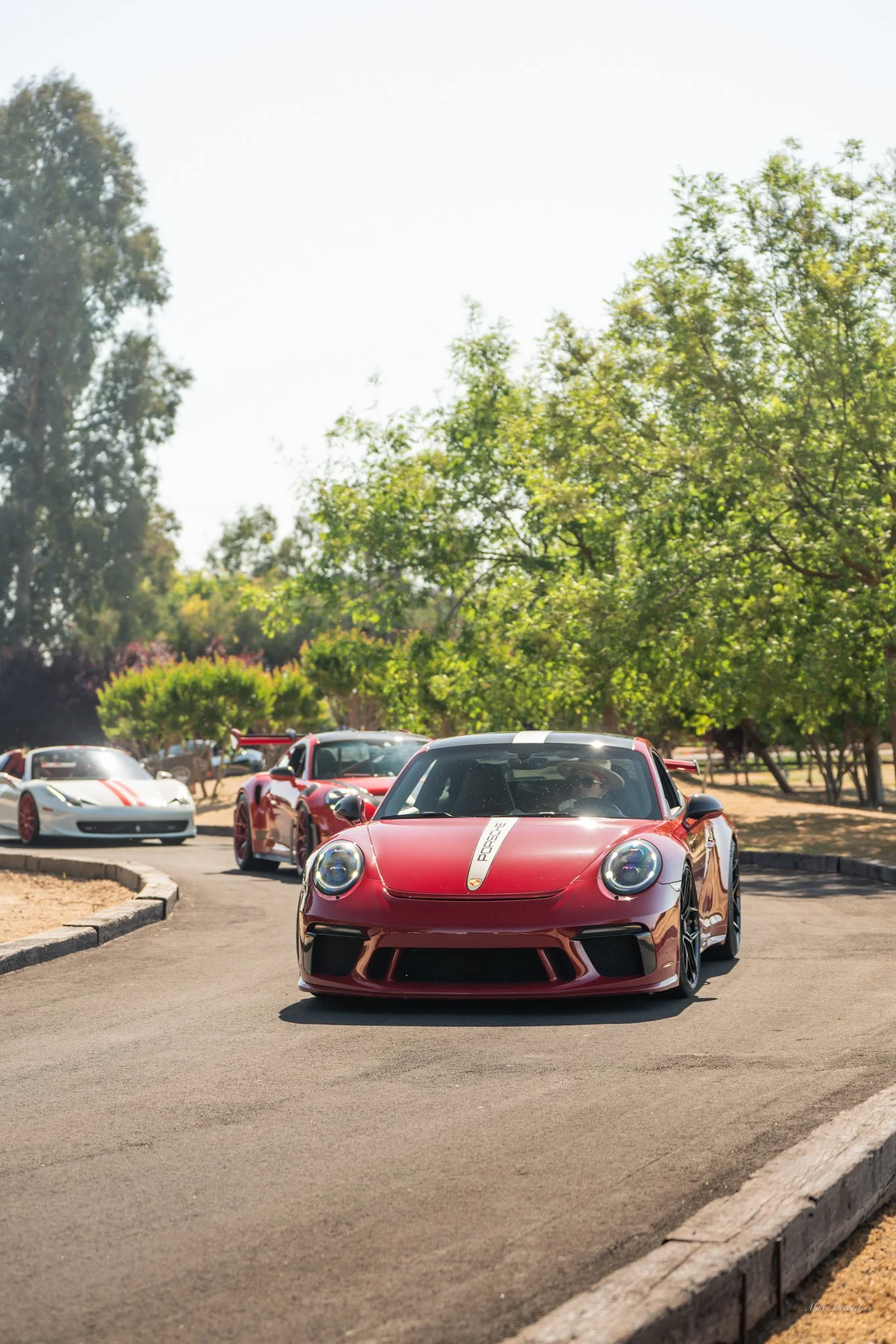 A red Porsche sports car with a white stripe driving on a curved road, followed by other luxury sports cars, in a park-like setting with trees and bright sky.