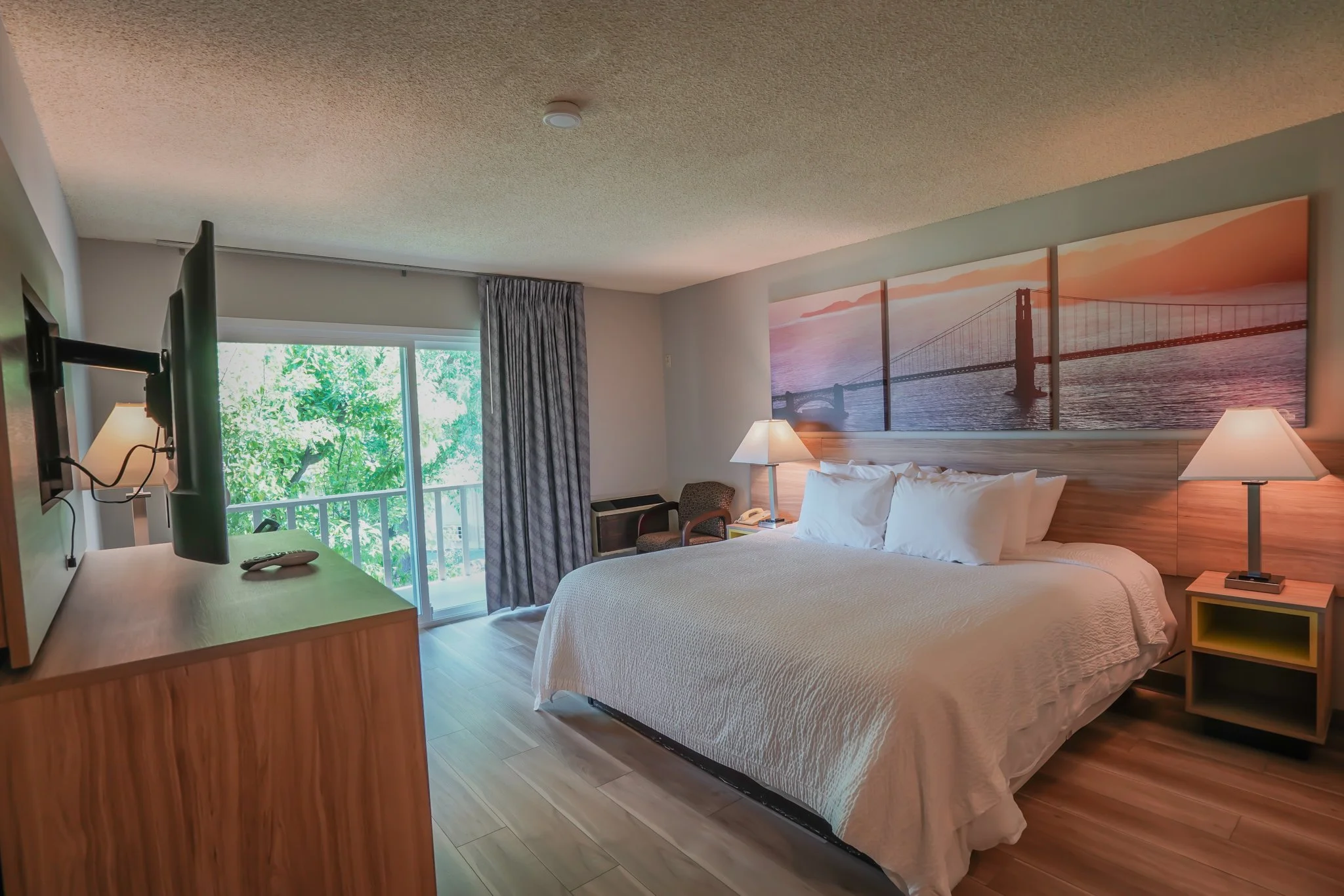 Hotel room with a large bed, nightstands with lamps, a seating chair, a sliding glass door with balcony, and a large mural of a bridge at sunset.