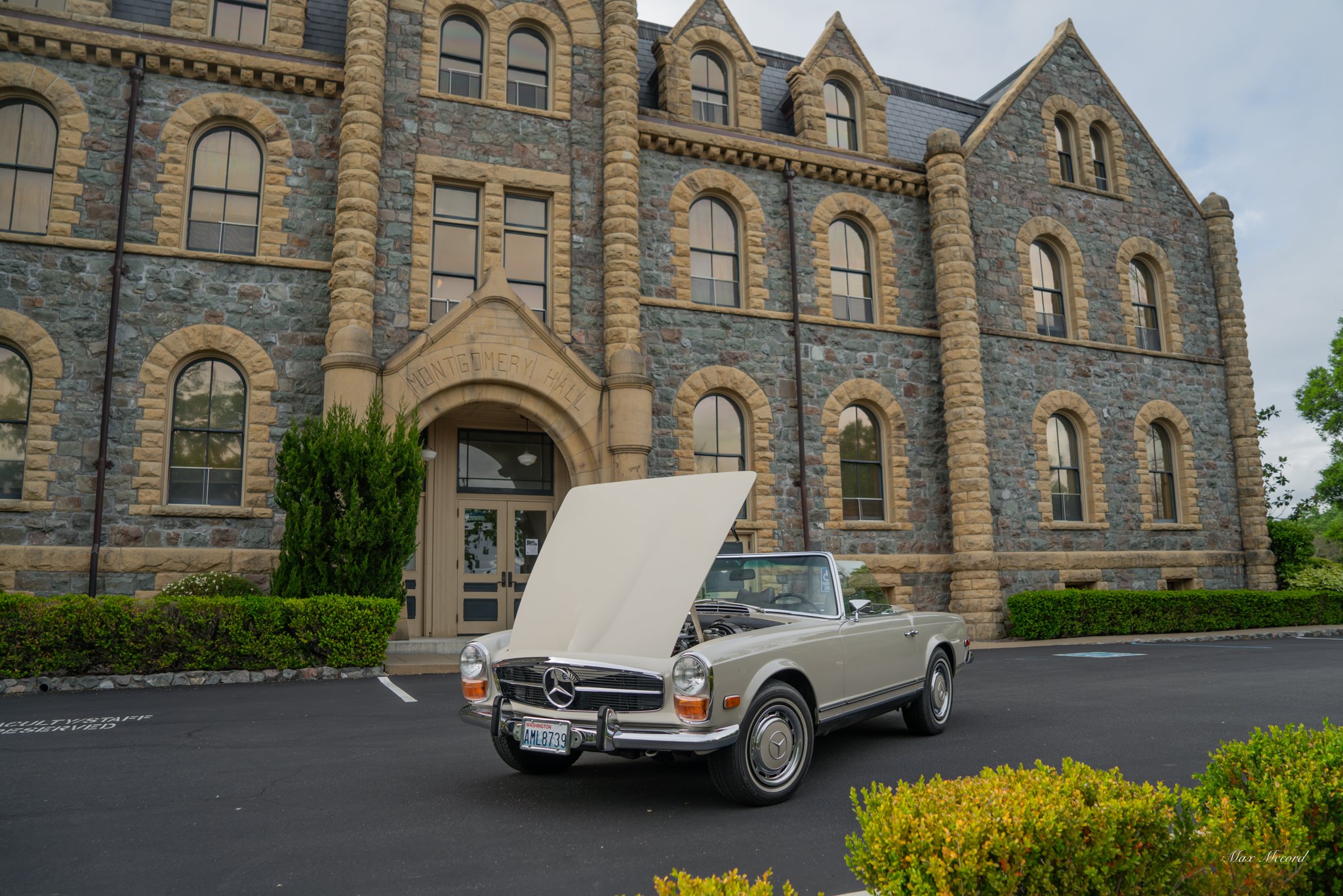 A vintage beige Mercedes-Benz convertible with its hood open, parked in front of Montgomery Hall, a stone building with arched windows and a decorative entrance, on a paved parking lot with green bushes and trees nearby.