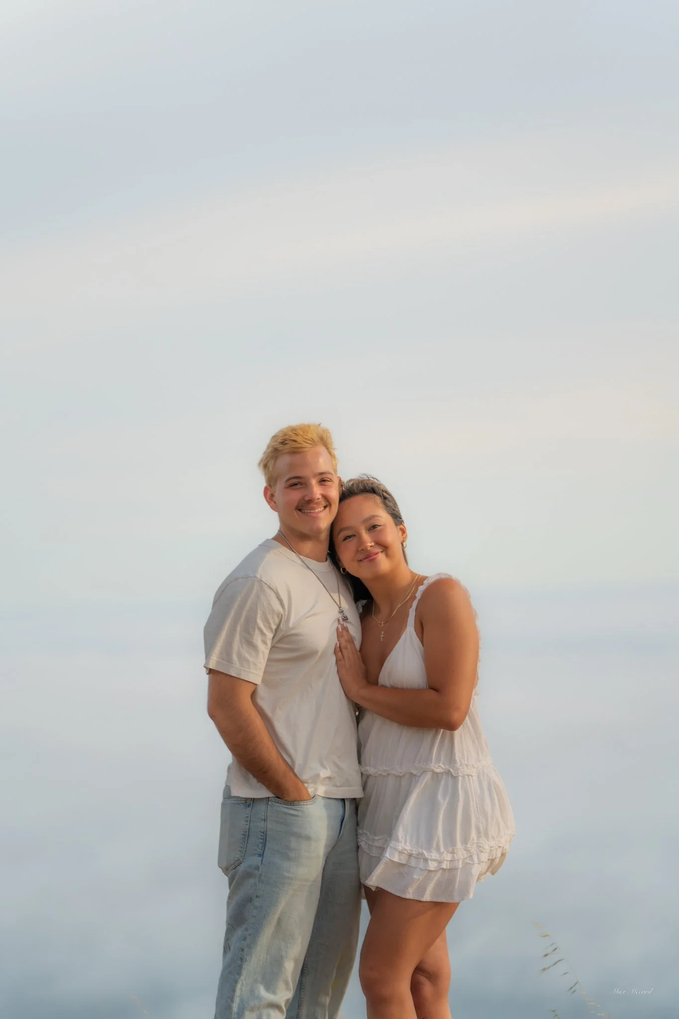 A young couple standing close together outdoors against a light sky, smiling at the camera. The man has blonde hair and is wearing a beige t-shirt and jeans. The woman has dark hair and is wearing a white dress, with her hand on the man's chest.