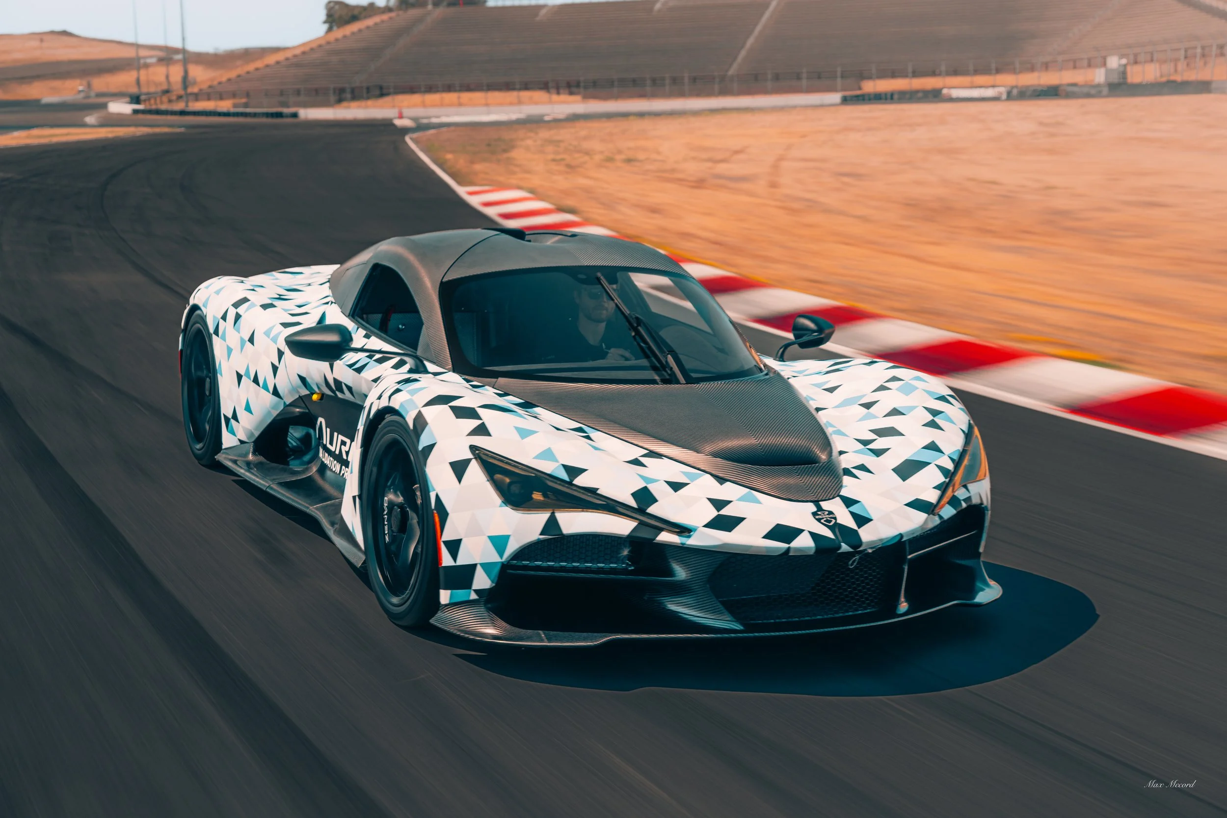 A race car with a black and white geometric pattern on the body and a black carbon fiber hood, driving on a race track with red and white curb markings and desert landscape in the background.