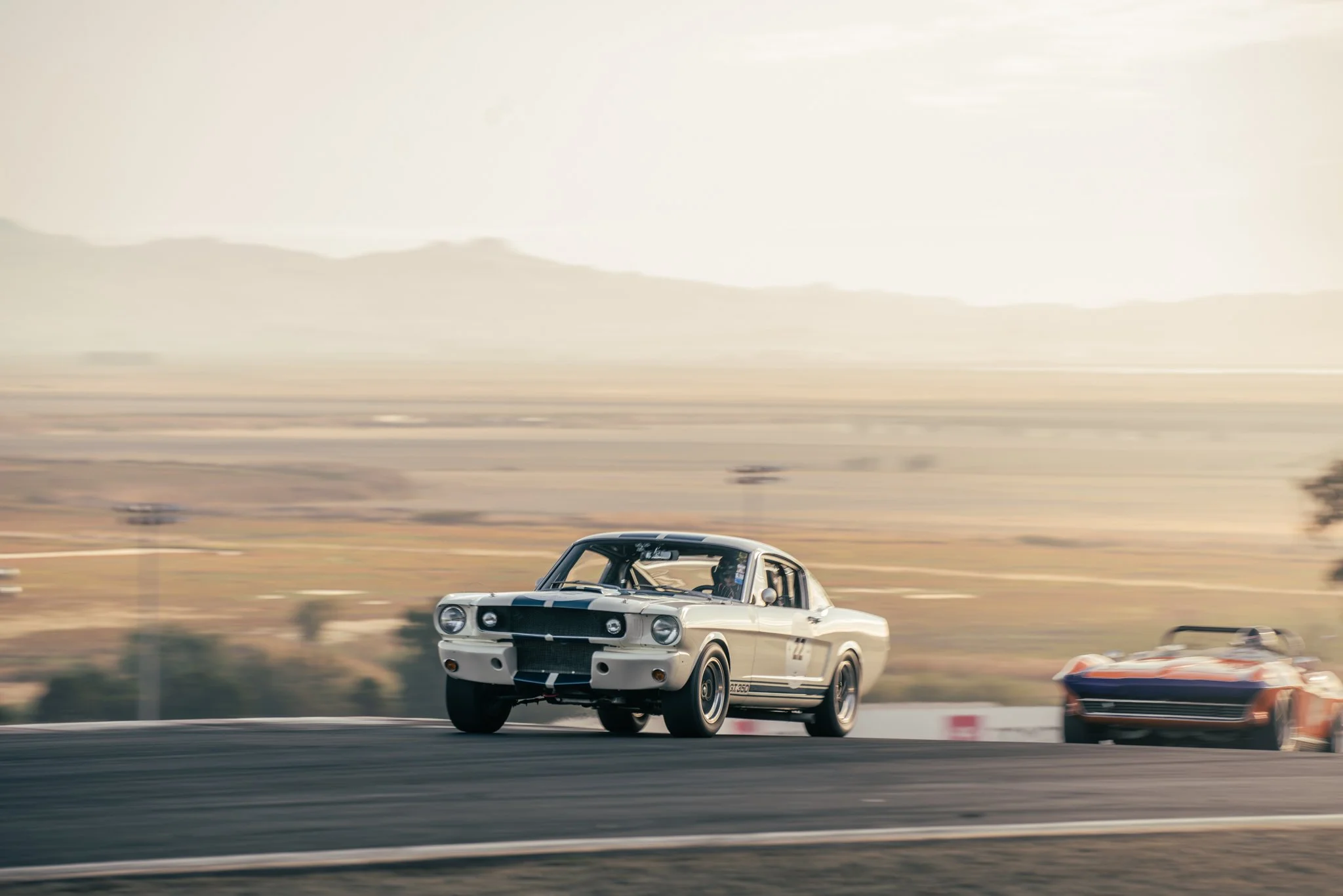 A vintage race car on a track with a second race car behind it, in a landscape with mountains in the distance during daylight.