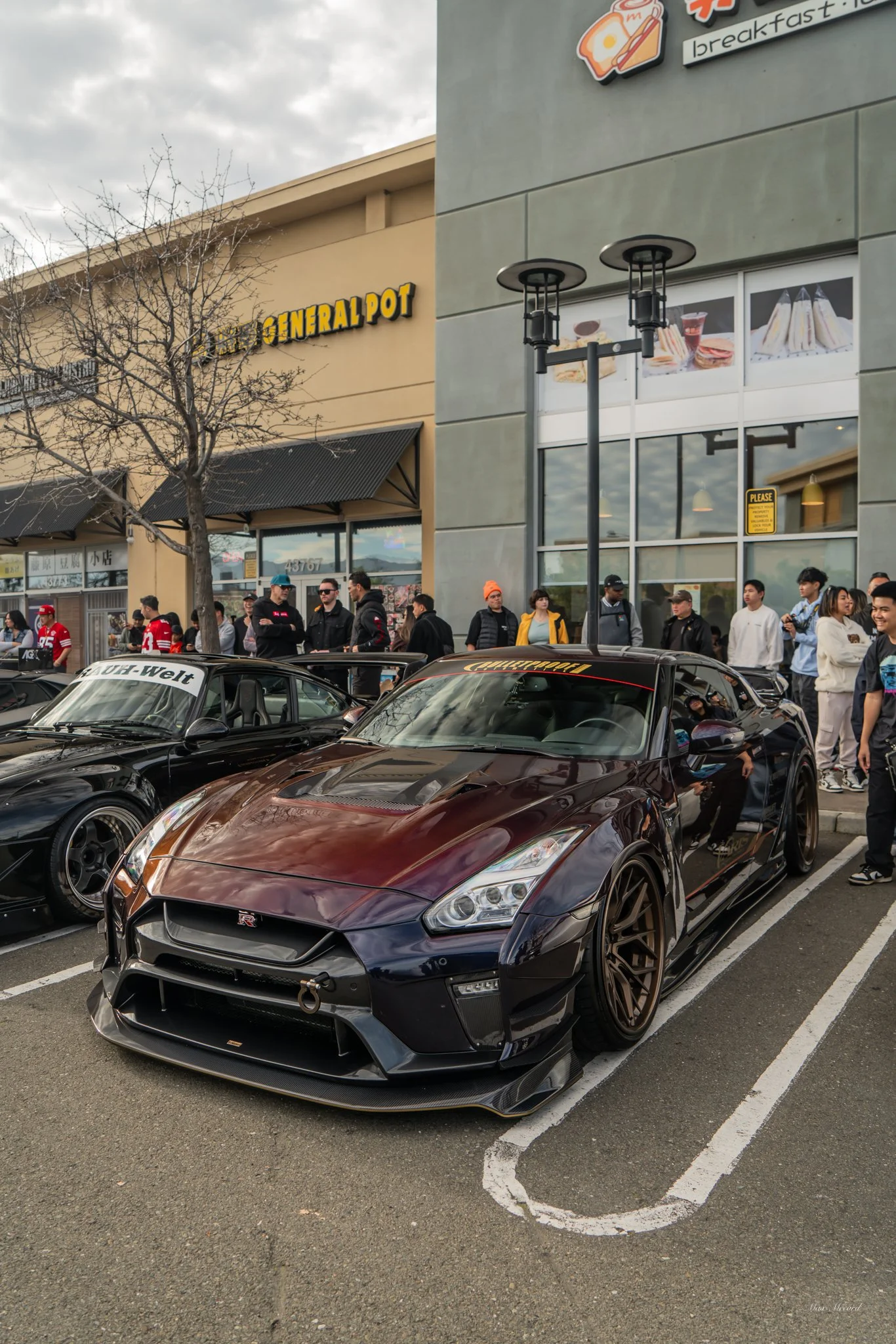A black Nissan GT-R sports car with a gradient paint job parked in a lot with a crowd of people around it and other cars beside it. In the background, there is a shopping center with a sign that says "The General Pot" and another sign showing breakfa