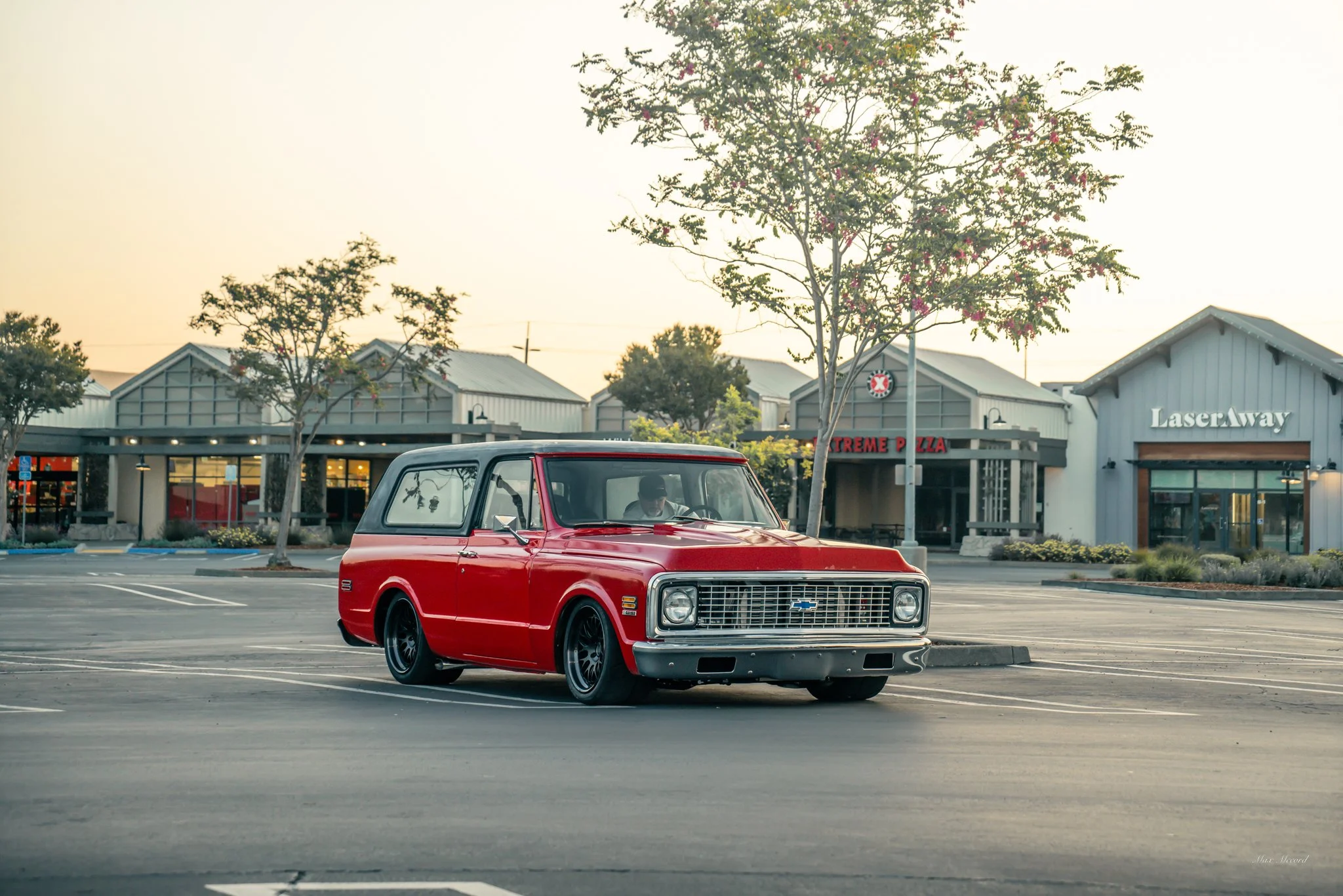 A red vintage Chevrolet station wagon parked in an empty parking lot during sunset, with a shopping center in the background and trees lining the area.