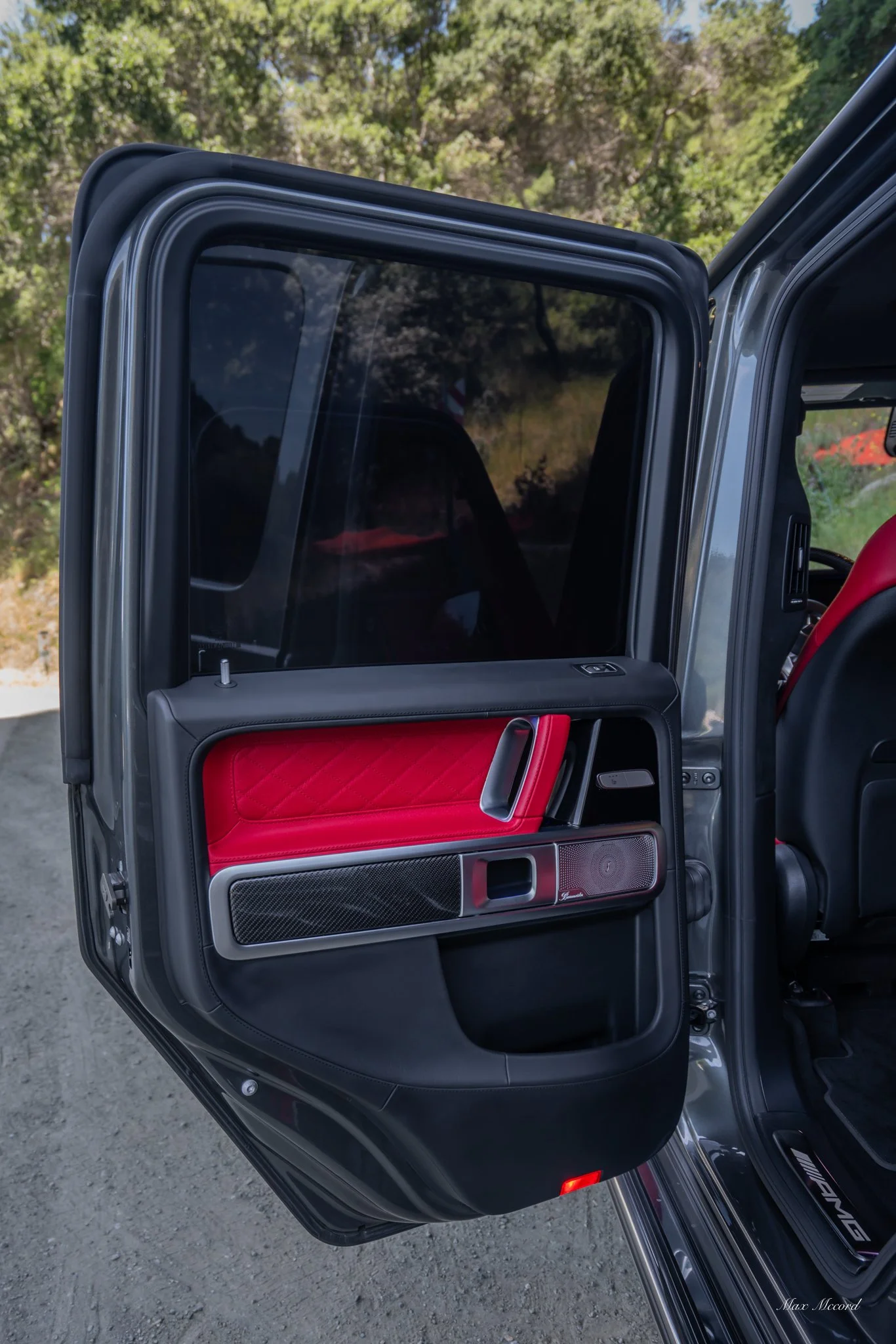 Open car door showing red and black interior with a carbon fiber speaker and handle, outdoors with trees in the background.