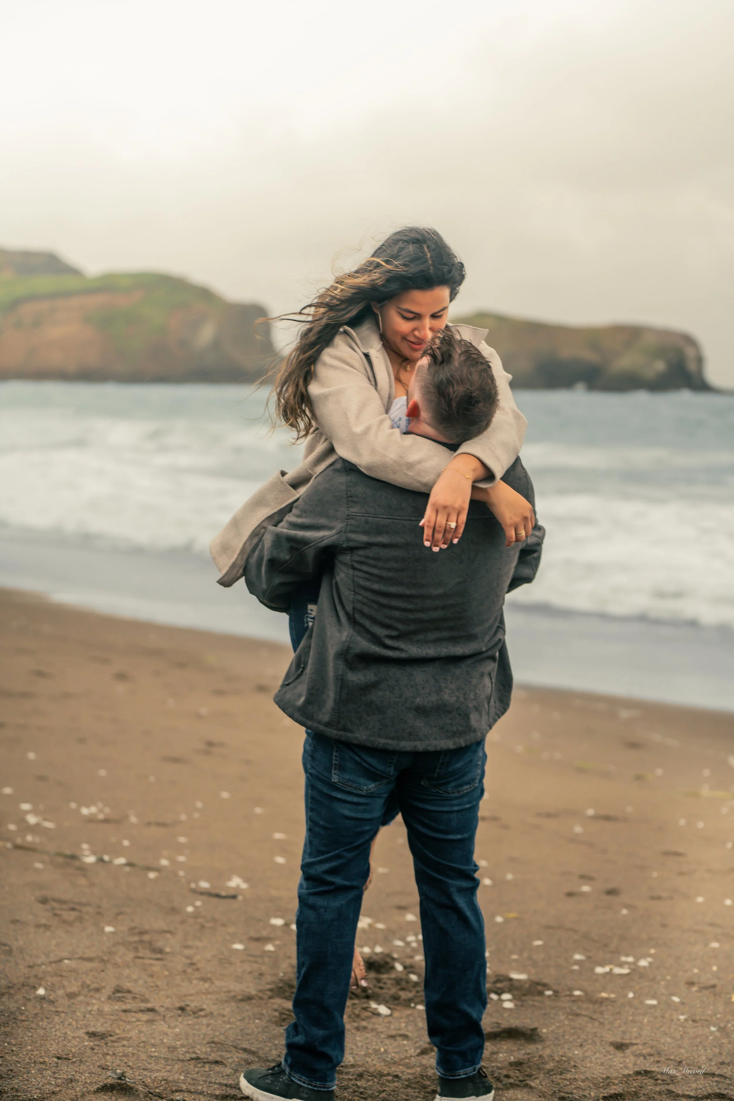 A woman being lifted into the air by a man on a beach, with waves and cliffs in the background.