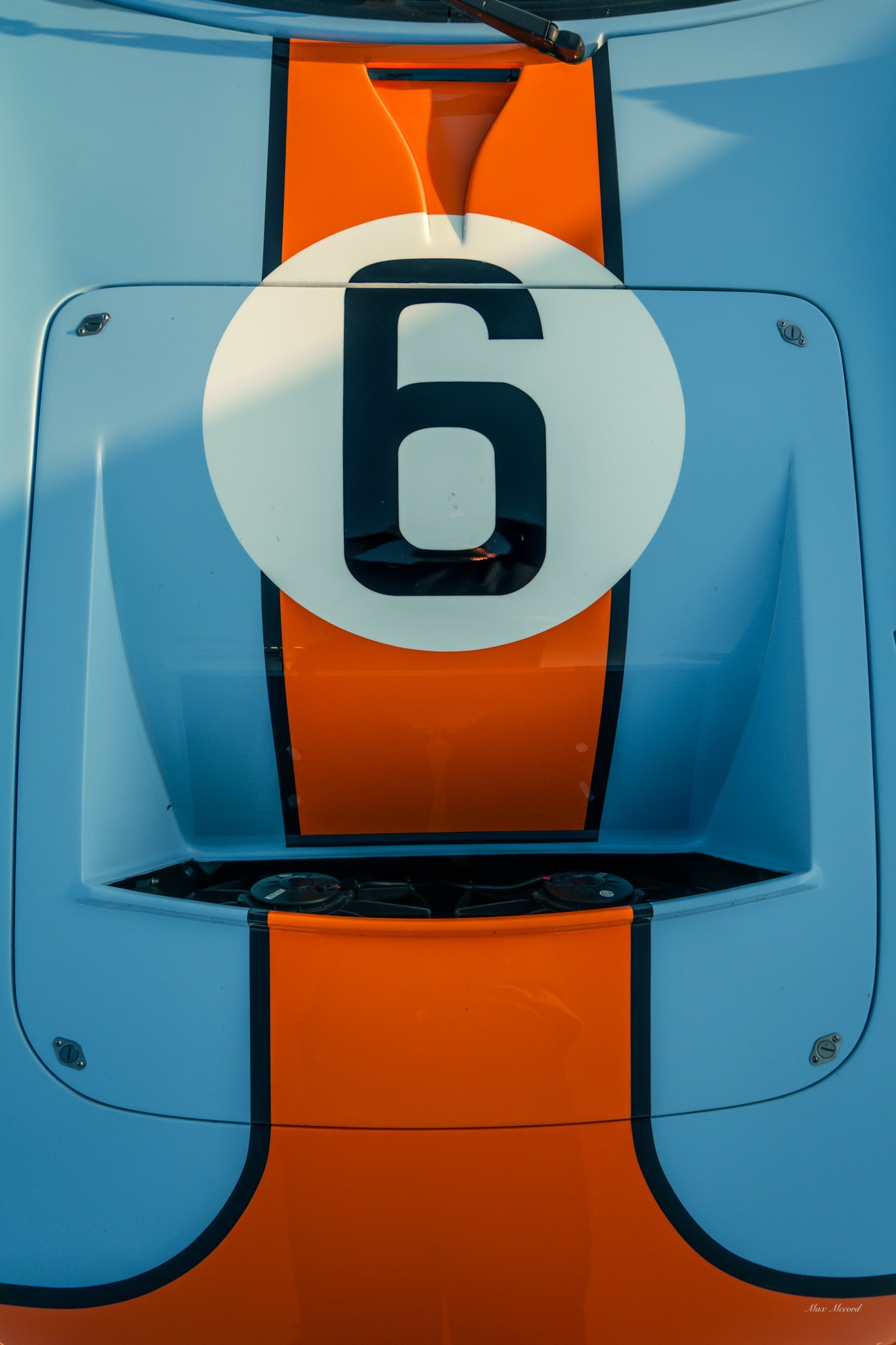 Close-up of a vintage racing car with the number 6 on a white circle on the hood, featuring orange and light blue colors and a sleek aerodynamic design.