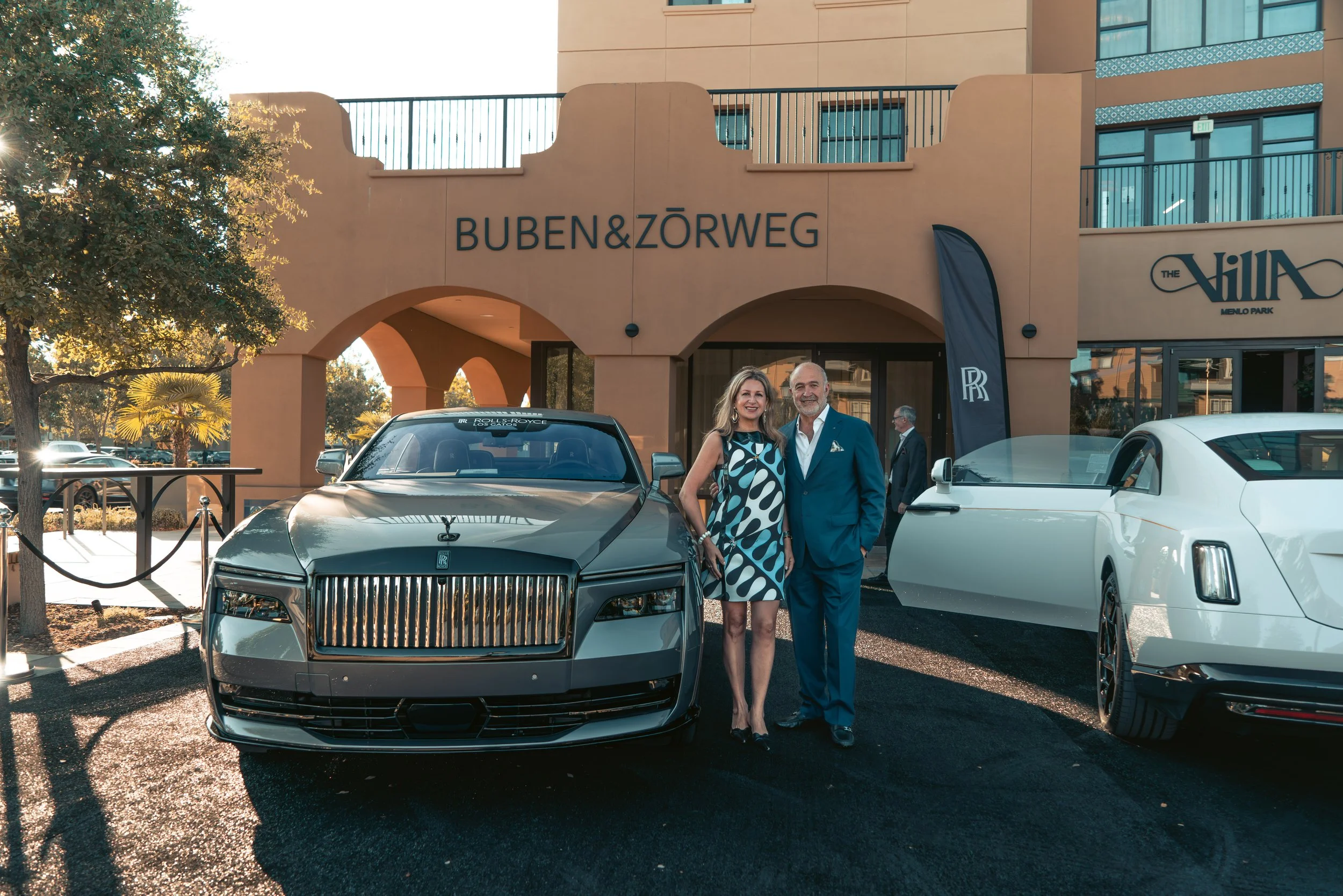 A man and woman dressed in formal attire standing in front of luxury cars outside a building with the sign BUBEN & ZÖRWEG, with a Rolls Royce behind them and another white luxury vehicle beside them.