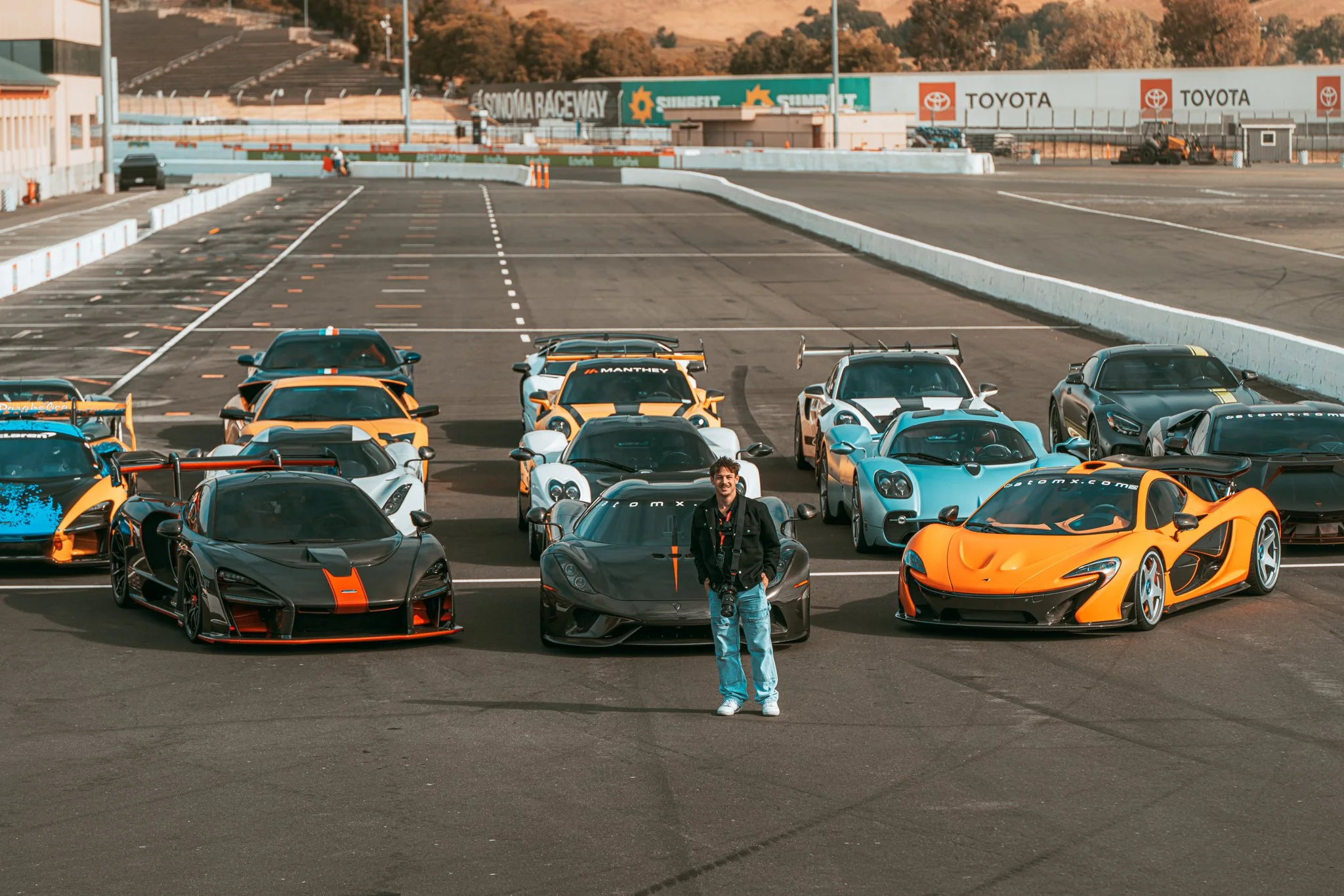 A person standing in front of a group of sports cars on a racetrack with a grandstand and signage in the background.