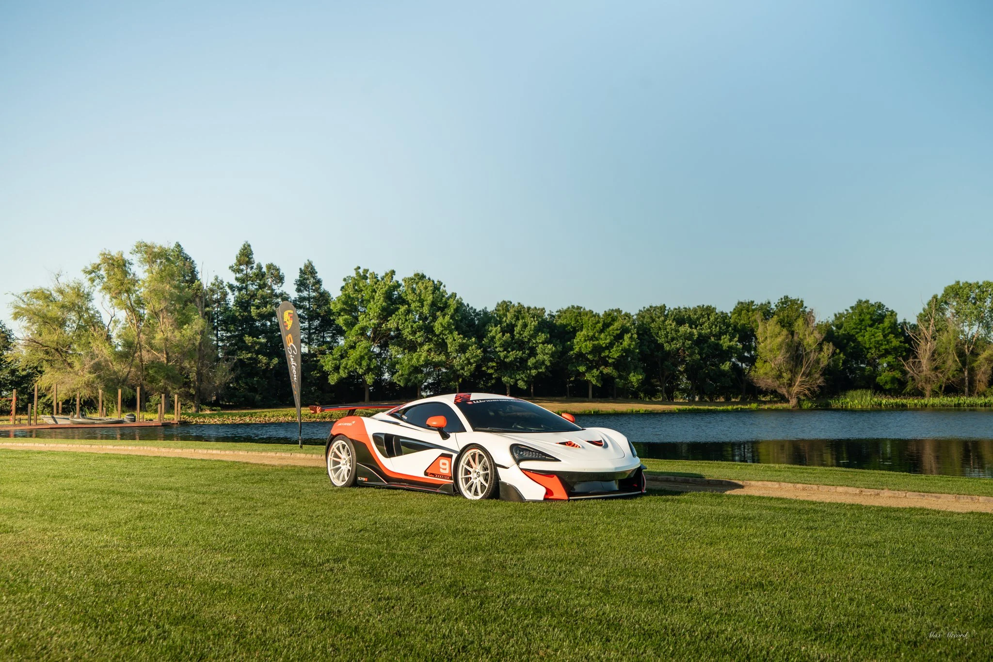 A white and black race car with orange accents parked on a grassy area near a lake with trees in the background.