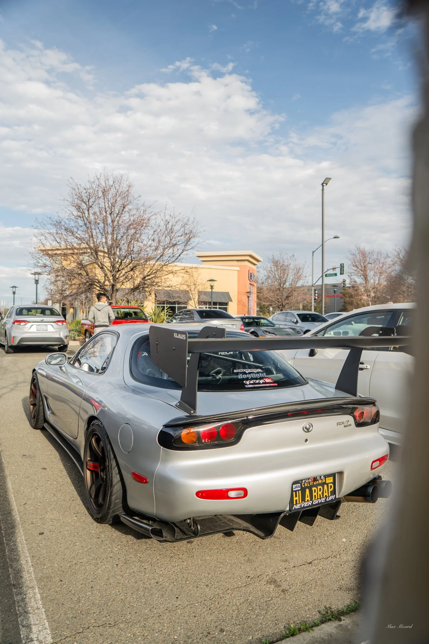 Rear view of a silver Mazda RX-7 sports car with a large rear wing in a parking lot, with several other cars and a shopping plaza in the background.