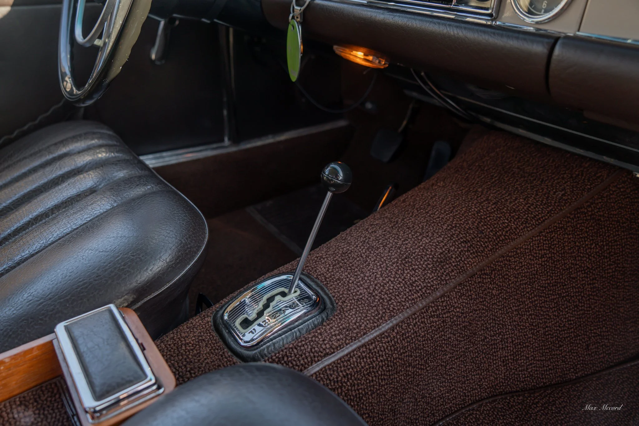 Close-up of a vintage car interior showing the gear shift with a black knob, brown textured upholstery, and parts of the dashboard and black leather seat.