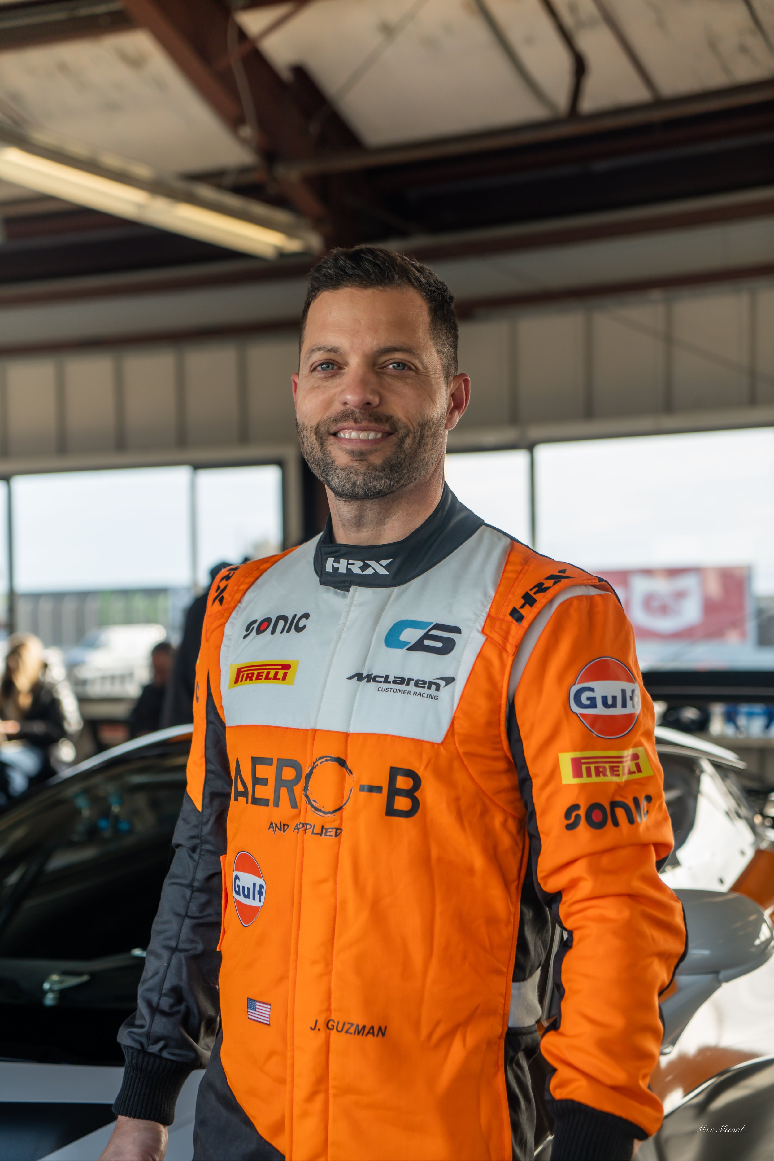 Male race car driver in orange racing suit smiling, standing in a garage with car and people in the background.