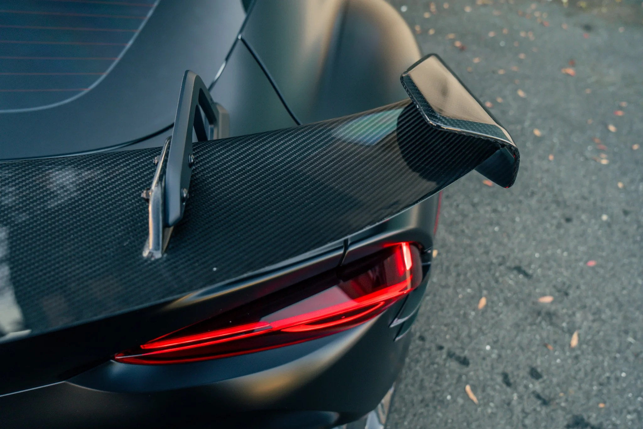 Close-up of the rear end of a sports car with a carbon fiber rear wing, sleek black body, and red tail lights on an asphalt road with small fallen leaves
