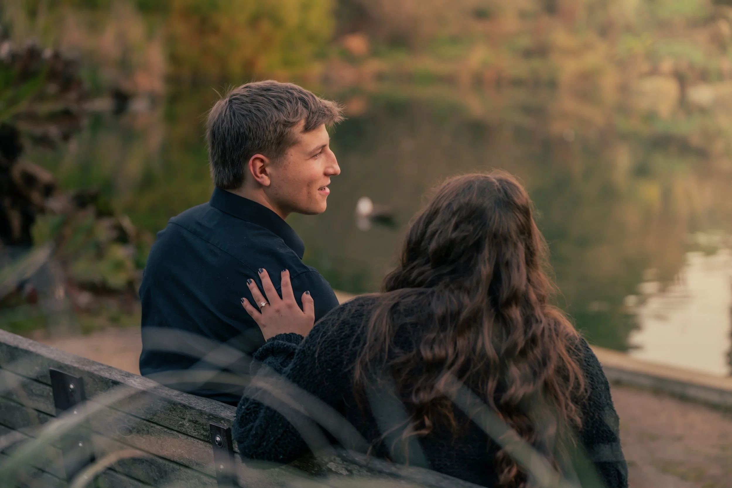 A young man and woman sitting on a park bench near a lake, surrounded by autumn trees, enjoying a peaceful moment together.