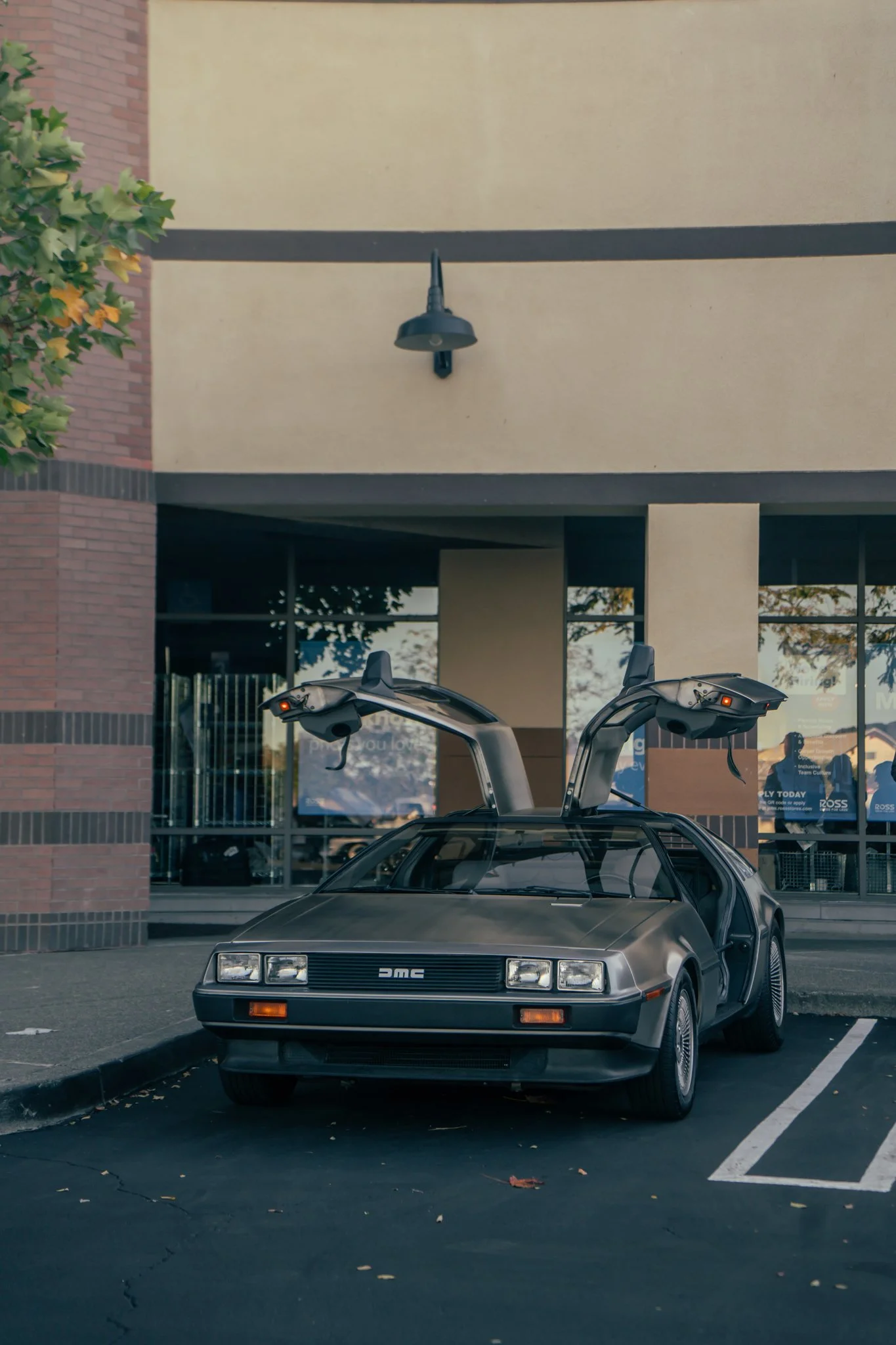A DeLorean DMC car with gull-wing doors open, parked in an outdoor lot in front of a building with large windows.