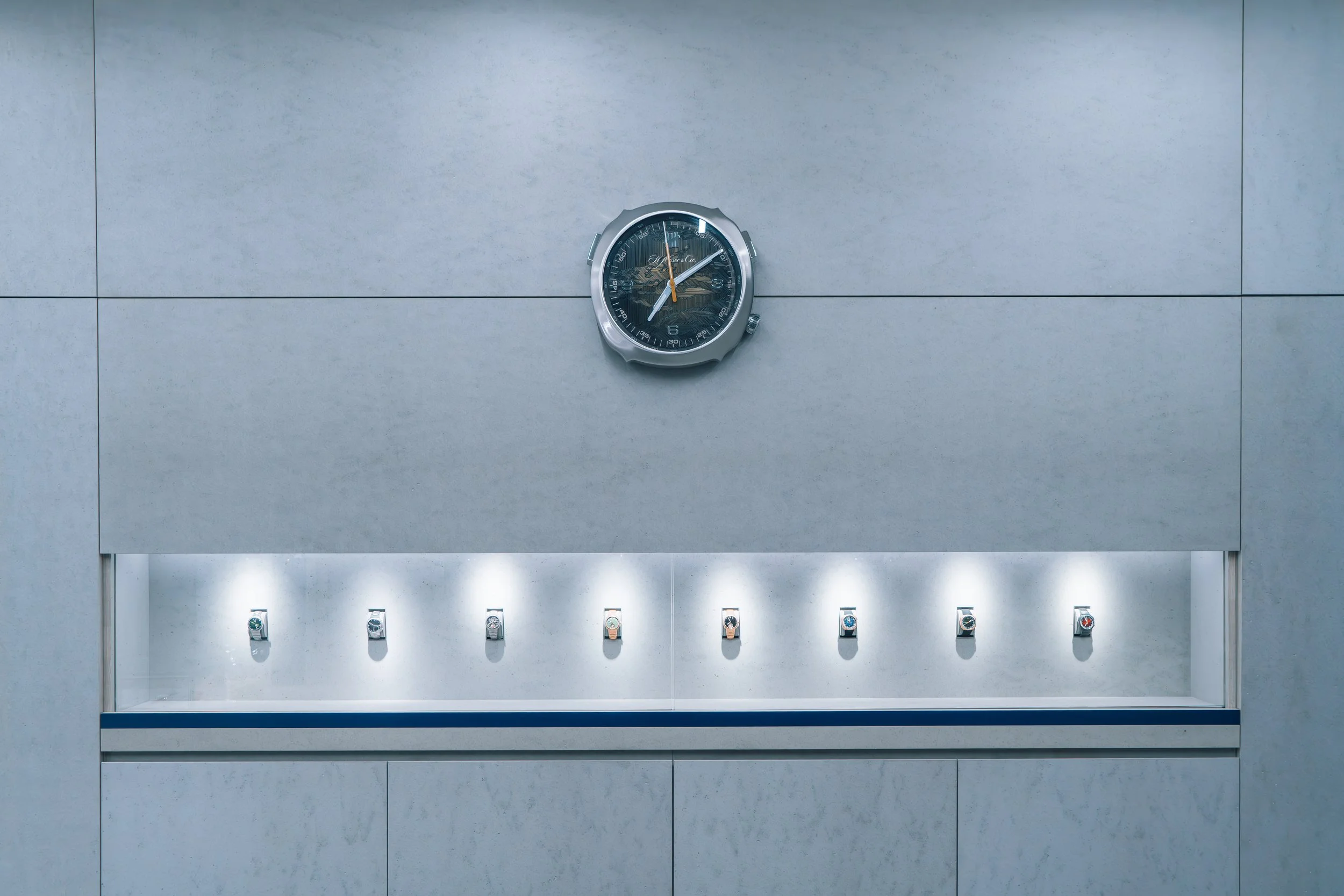 Display case of eight wristwatches and a wall clock above in an indoor setting.