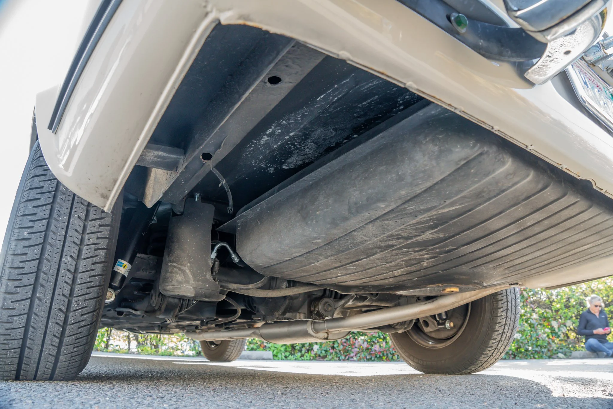 Underneath view of a car showing the fuel tank, suspension, and exhaust system on a paved surface, with a woman sitting in a background on the right side.