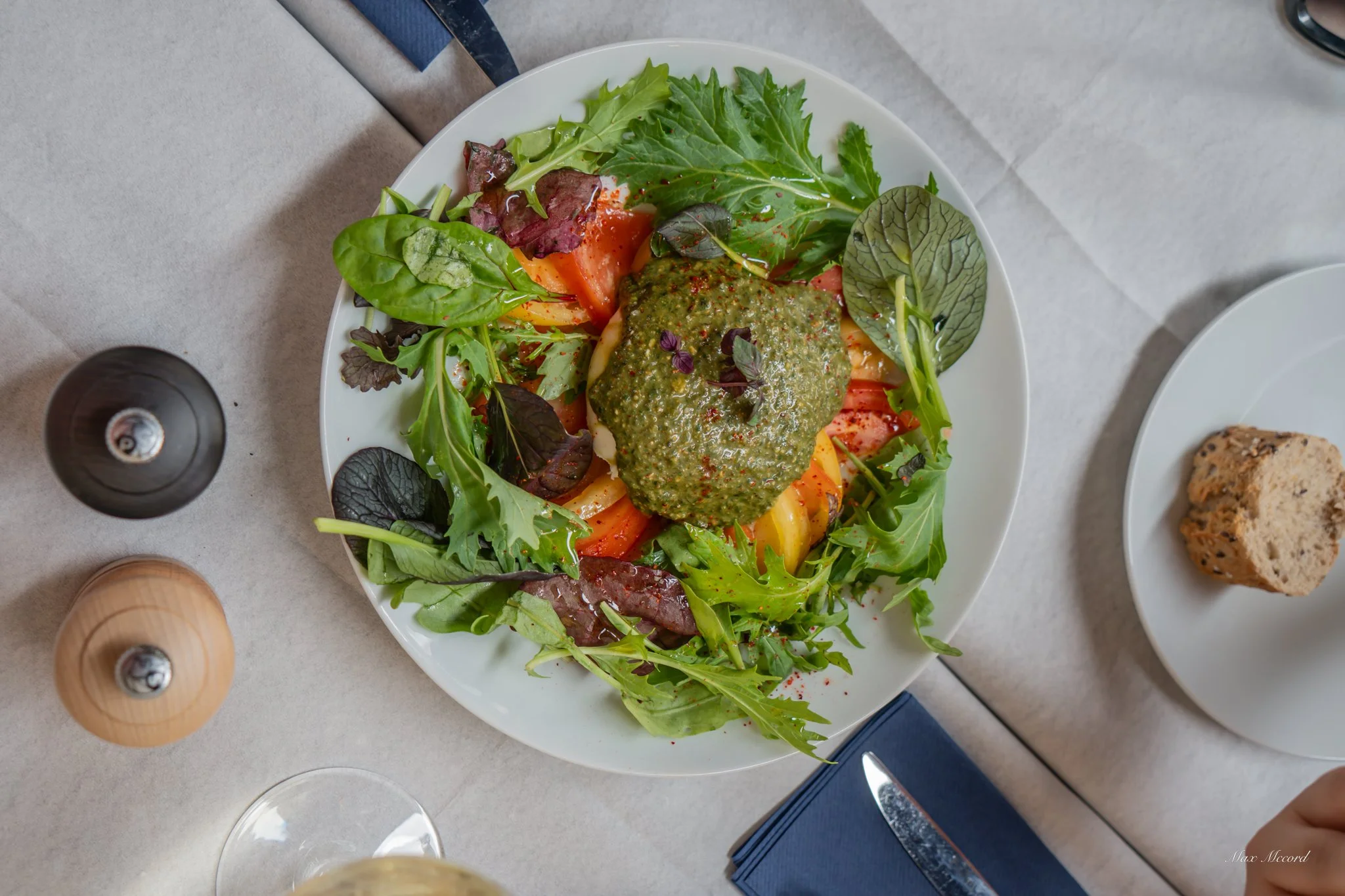 Fresh mixed green salad with tomato slices and a dollop of green pesto on top, served on a white plate. There is a piece of bread on a small white plate nearby, on a light-colored table with salt and pepper shakers, a blue napkin, and a glass of wate