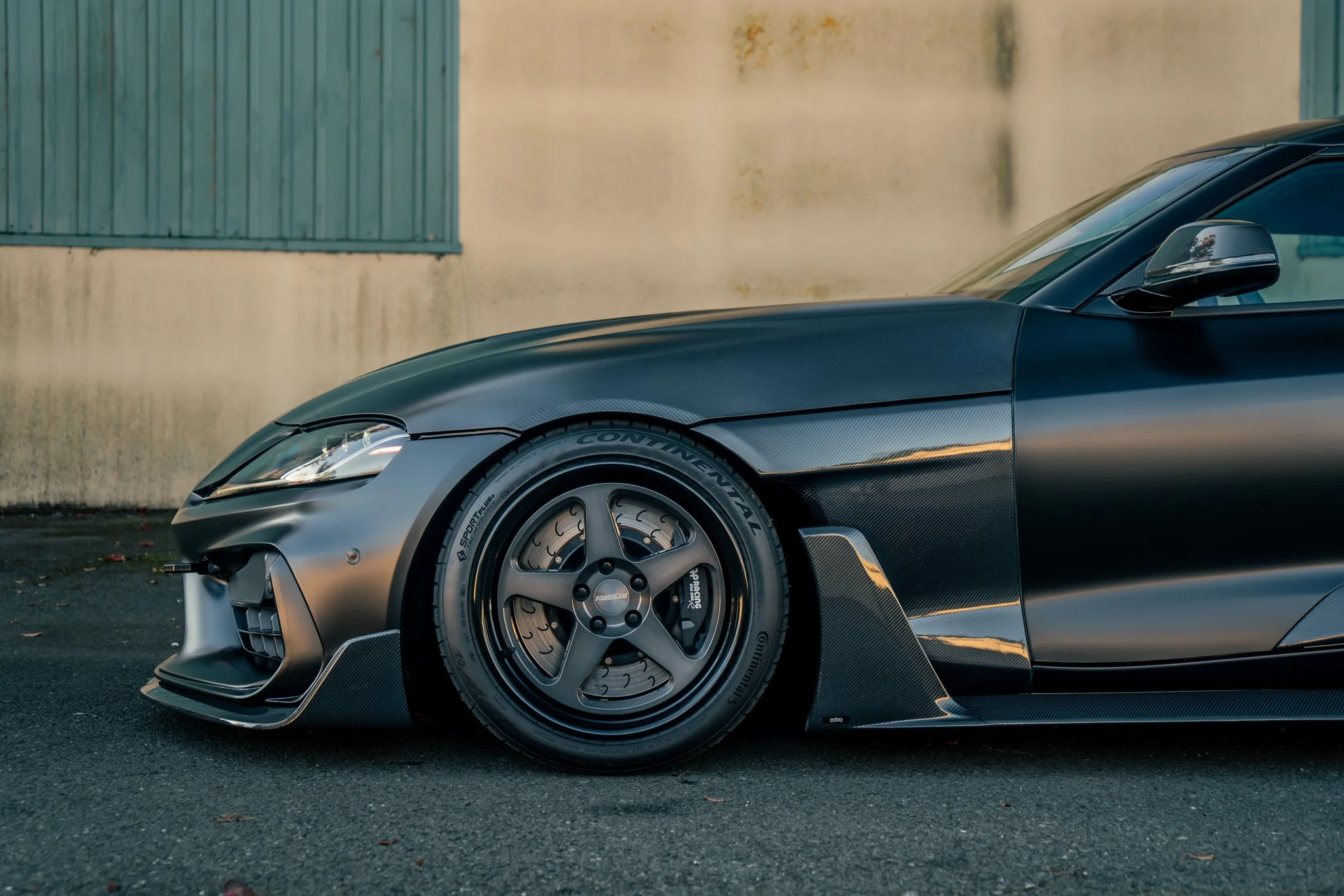 Close-up of a matte black sports car with carbon fiber accents, showing the front wheel and part of the side, parked on asphalt near a beige wall with a blue shutter.