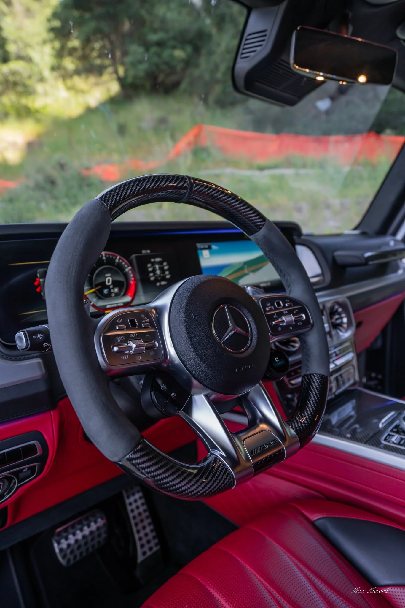 Interior of a luxury Mercedes-Benz vehicle with a red leather seat, a carbon fiber steering wheel, and a digital dashboard, parked outdoors near greenery.