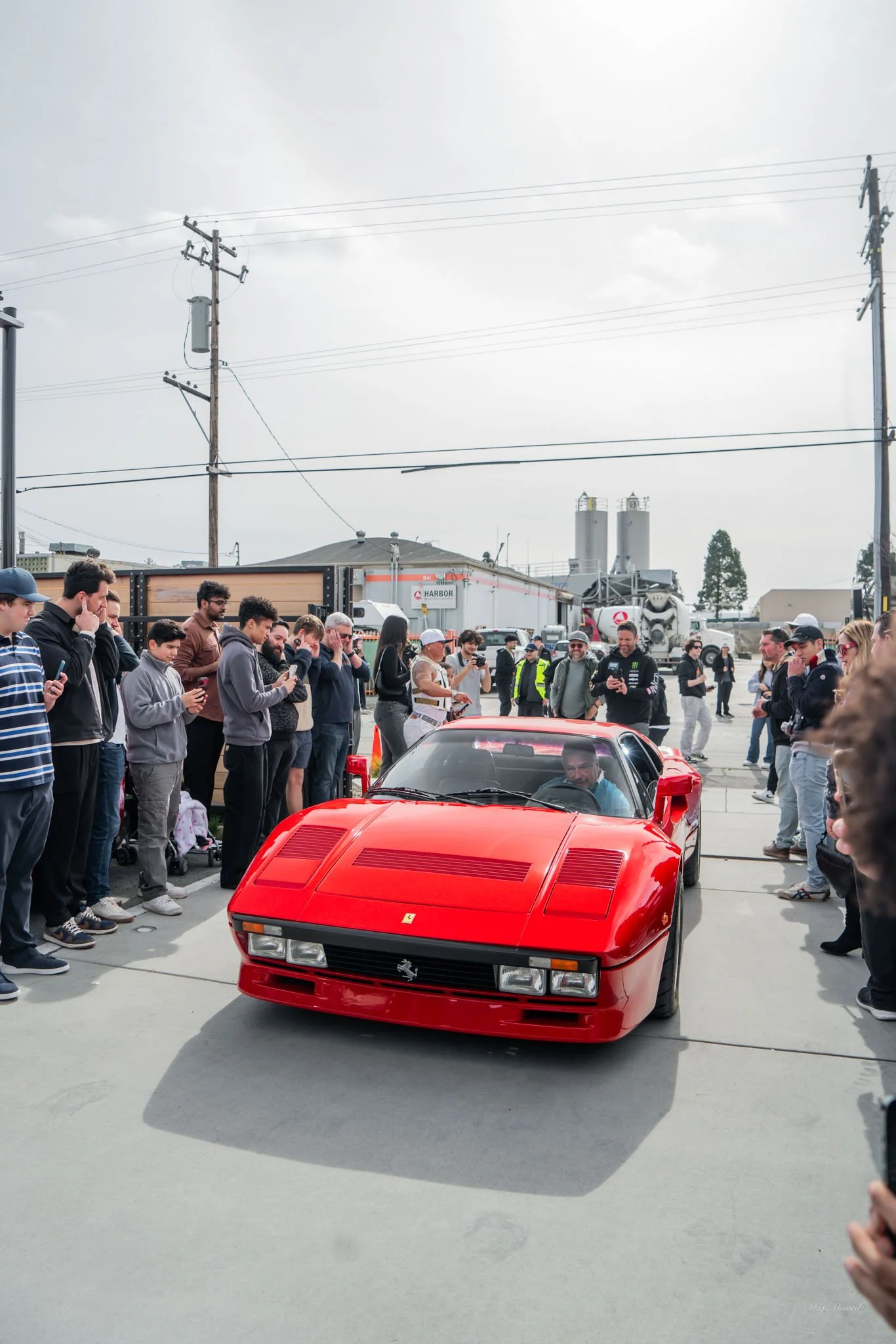 A red Ferrari sports car driving through a crowd of people taking photos and videos, outdoors on a cloudy day.