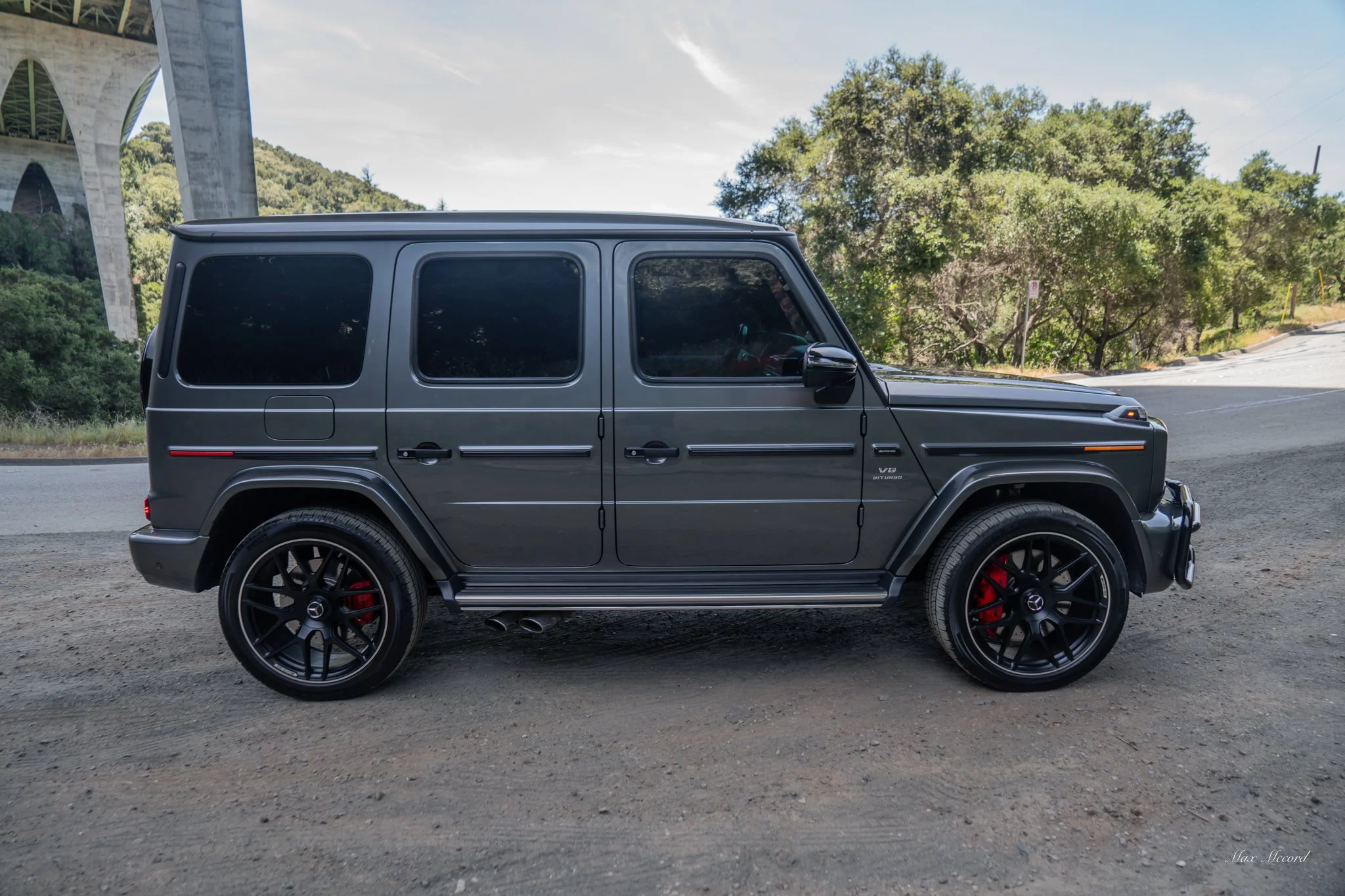 Side view of a gray Mercedes-Benz G-Class SUV parked on a dirt road with a bridge and trees in the background.