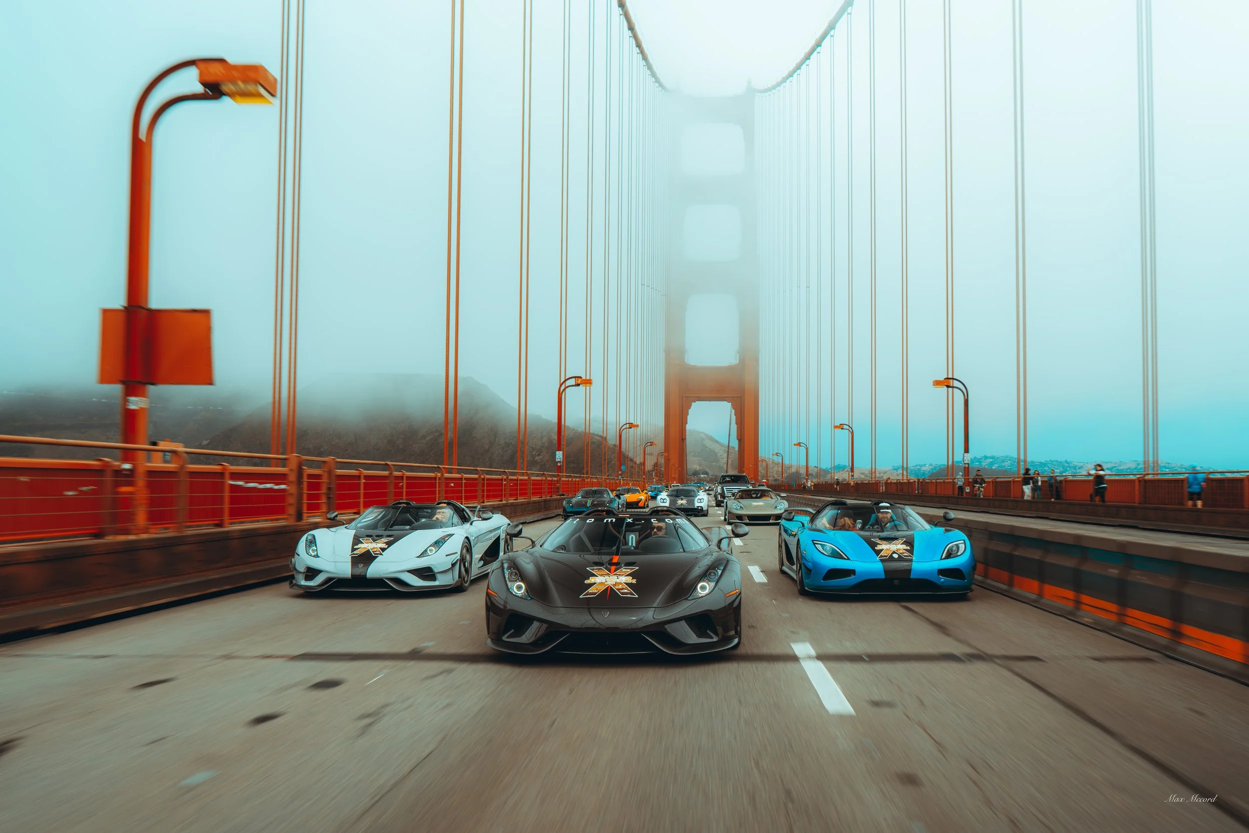 Multiple high-performance sports cars, including a black, blue, and white model, driving on the Golden Gate Bridge amidst foggy weather, with the bridge's towers and suspension cables visible.