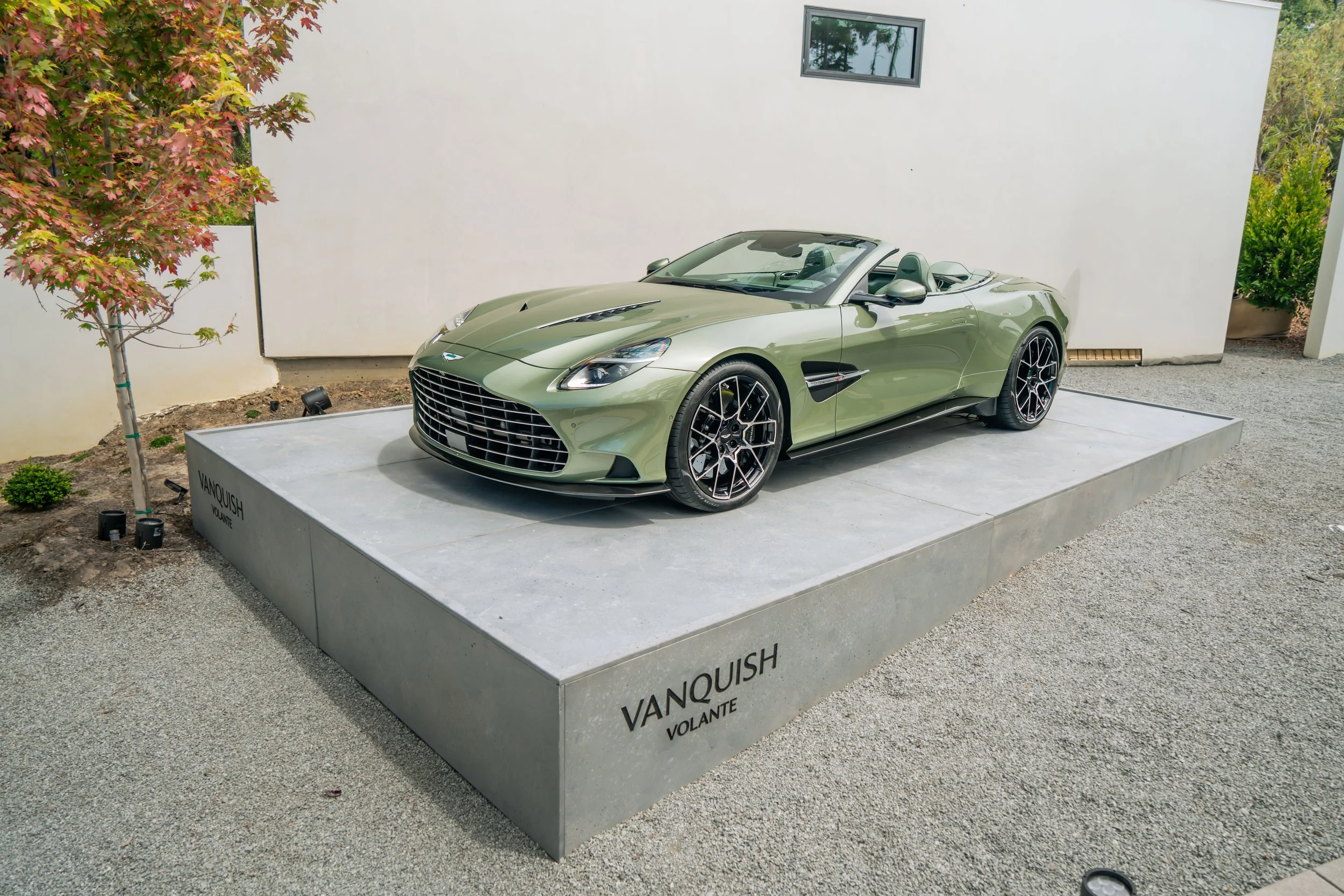A light green luxury convertible sports car displayed on a gray platform outdoors, labeled 'VANQUISH VOLANTE', with a white wall and small tree with red leaves in the background.