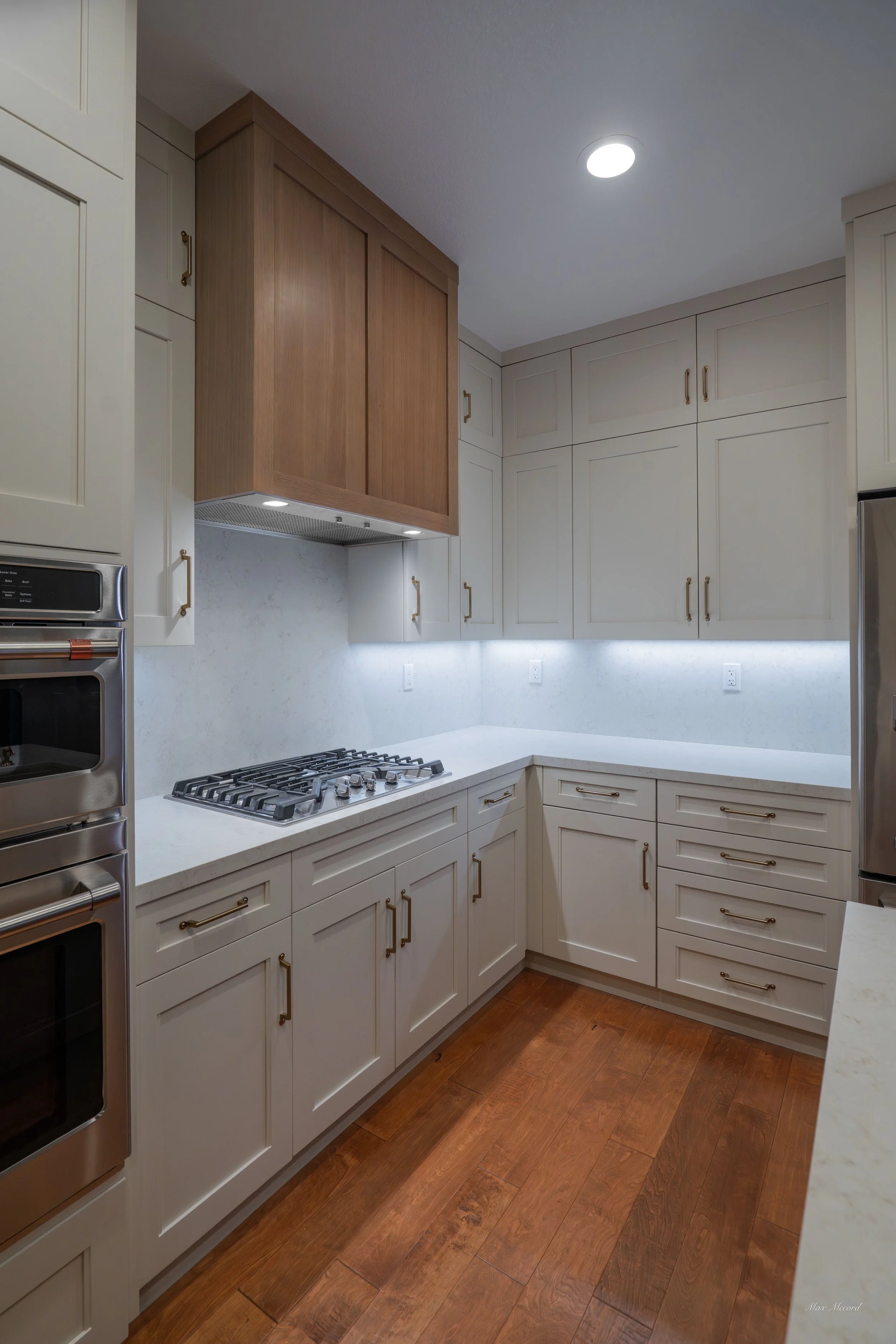Modern kitchen with white cabinets, wooden flooring, and stainless steel appliances, including a double wall oven, gas stovetop, and refrigerator.
