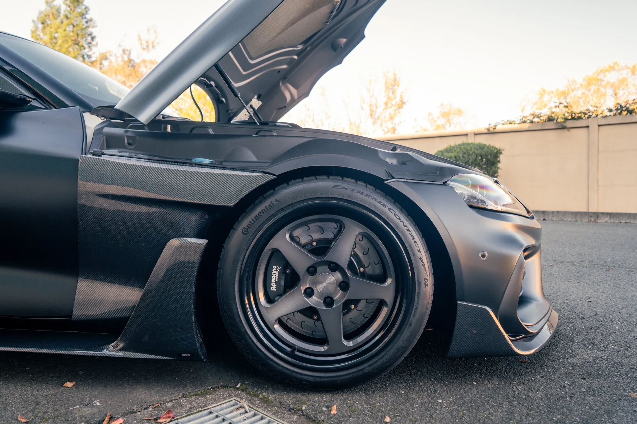 Close-up of a black sports car with an open hood, showing a large rear wheel with a racing tire and a carbon fiber side skirt.