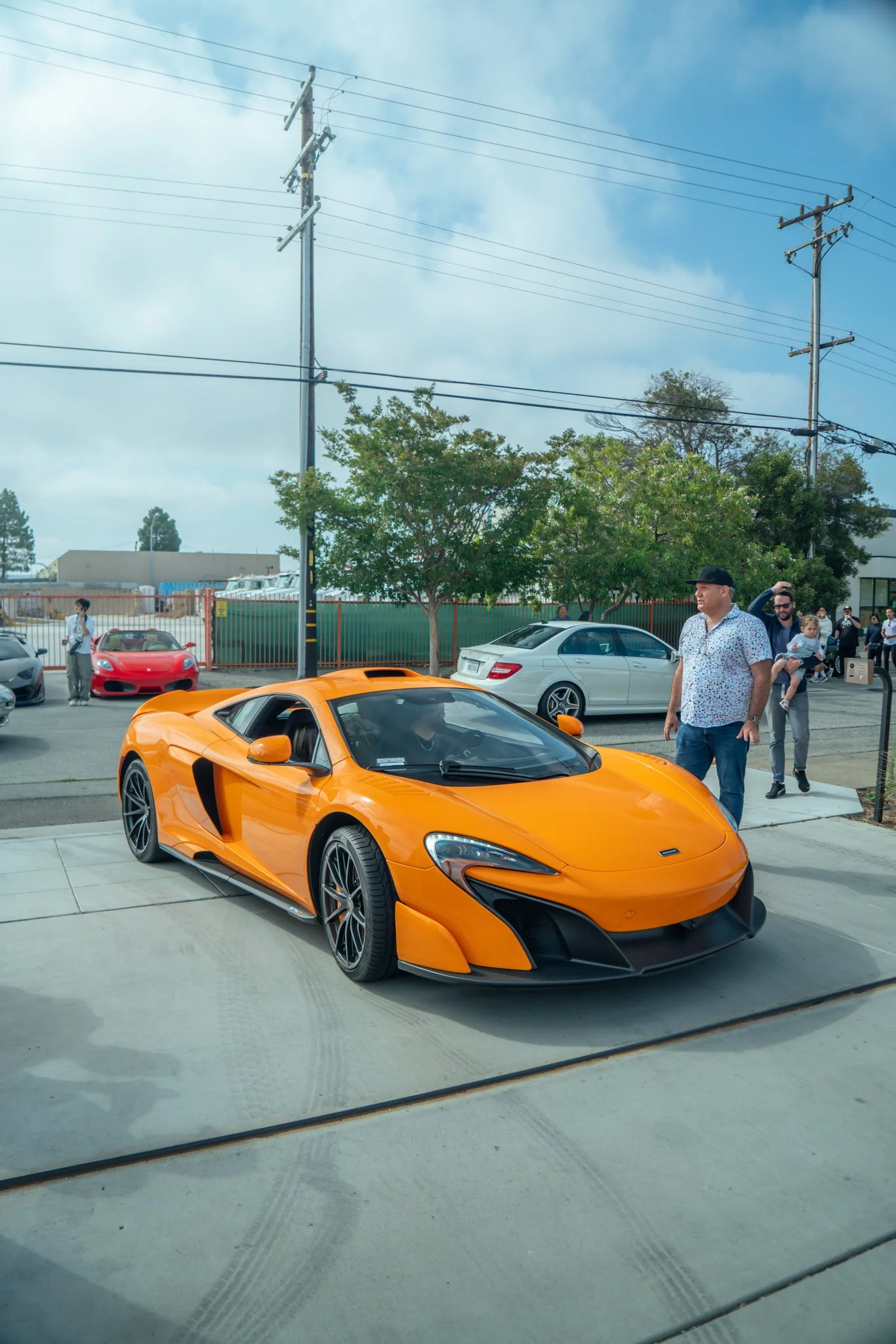 An orange supercar parked on a concrete sidewalk, with several people standing nearby, and trees, other cars, and a fence in the background.