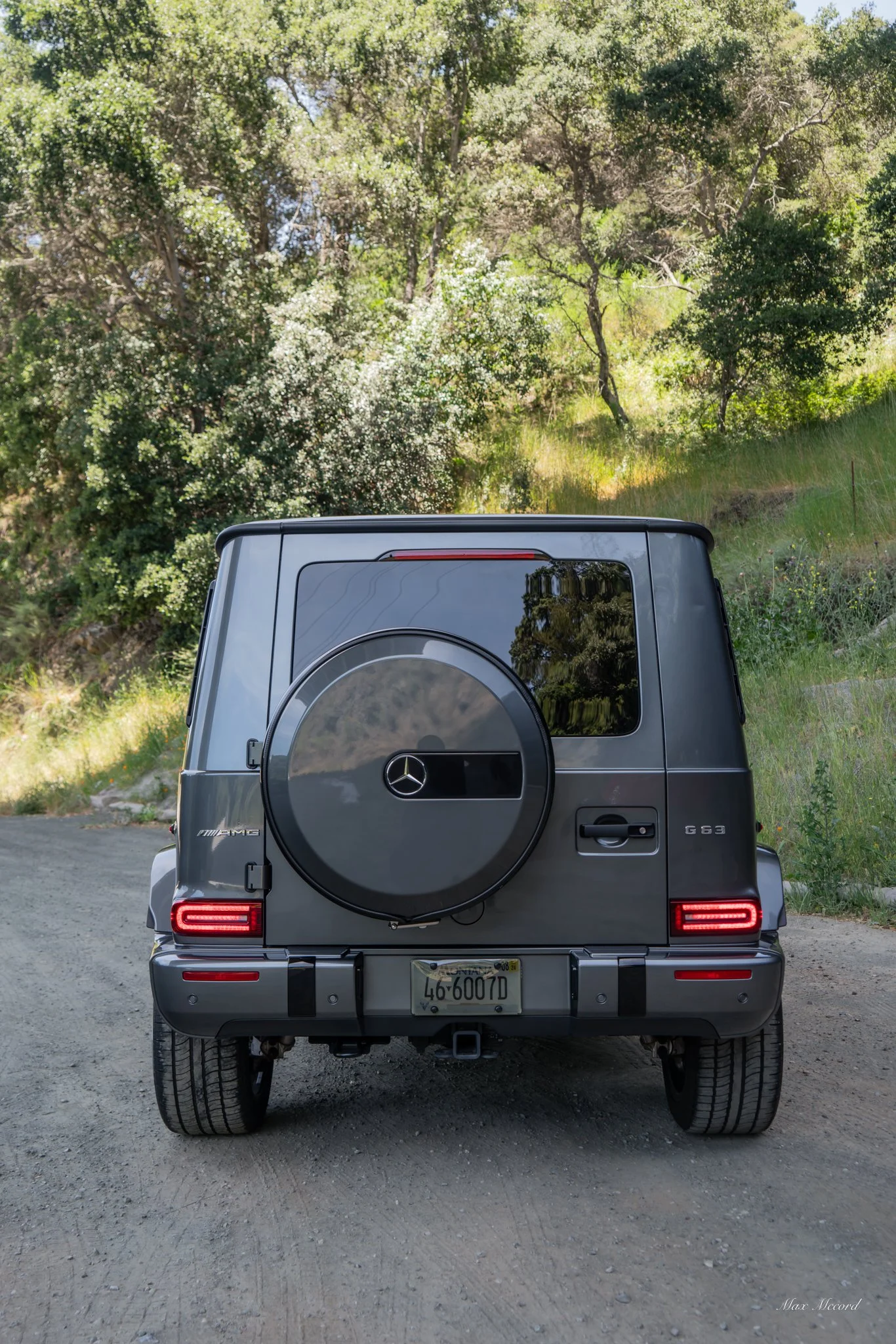 Rear view of a dark gray Mercedes-Benz G-Class SUV parked on a dirt road surrounded by green trees and foliage.