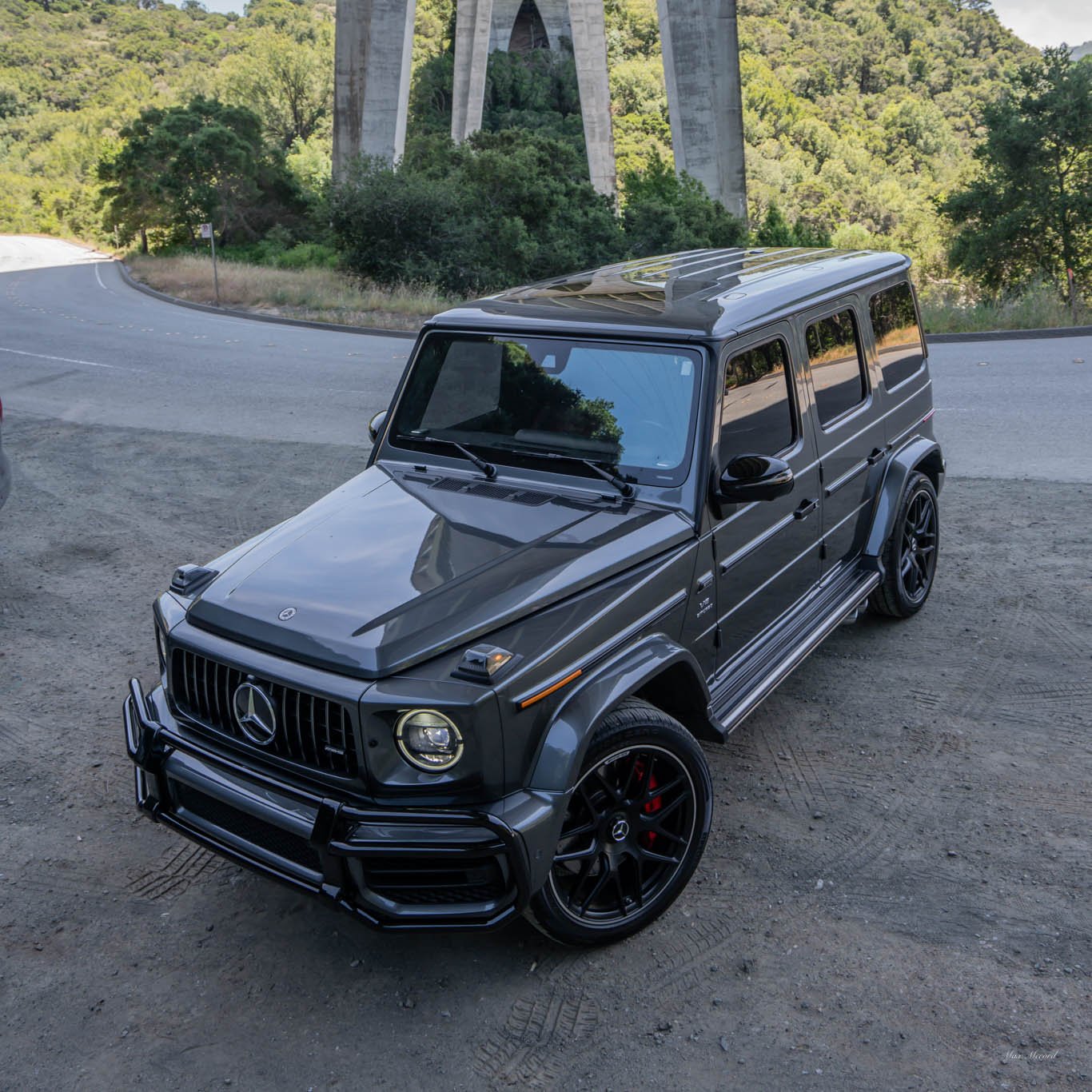 A black Mercedes-Benz G-Class SUV parked on a dirt area near a curved road with a bridge and green trees in the background.
