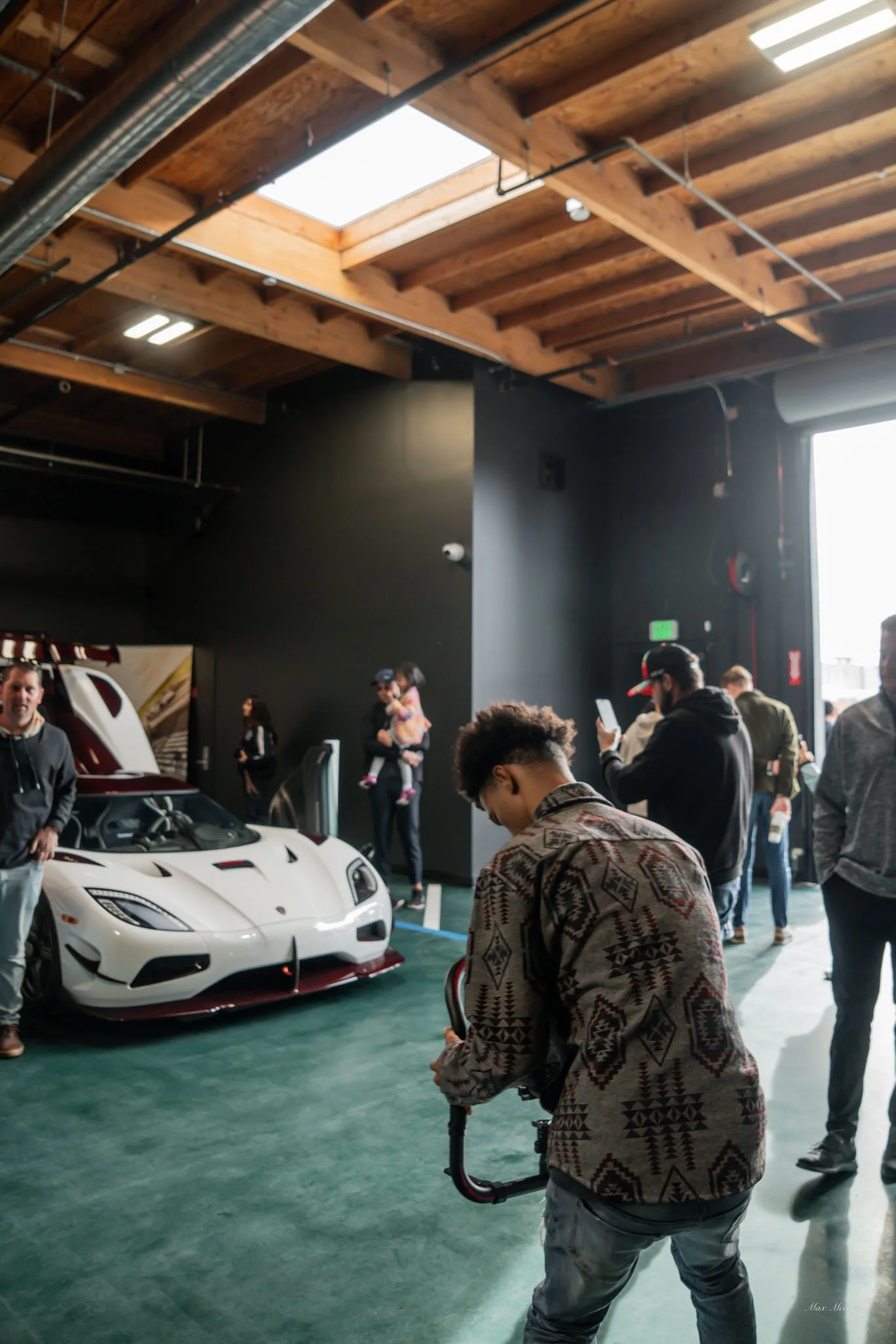 A white sports car with an open rear hatch is displayed indoors. Several people are nearby, some taking photos or talking. The setting features an industrial-style ceiling with wooden beams and skylights, bright natural light, and a black wall in the