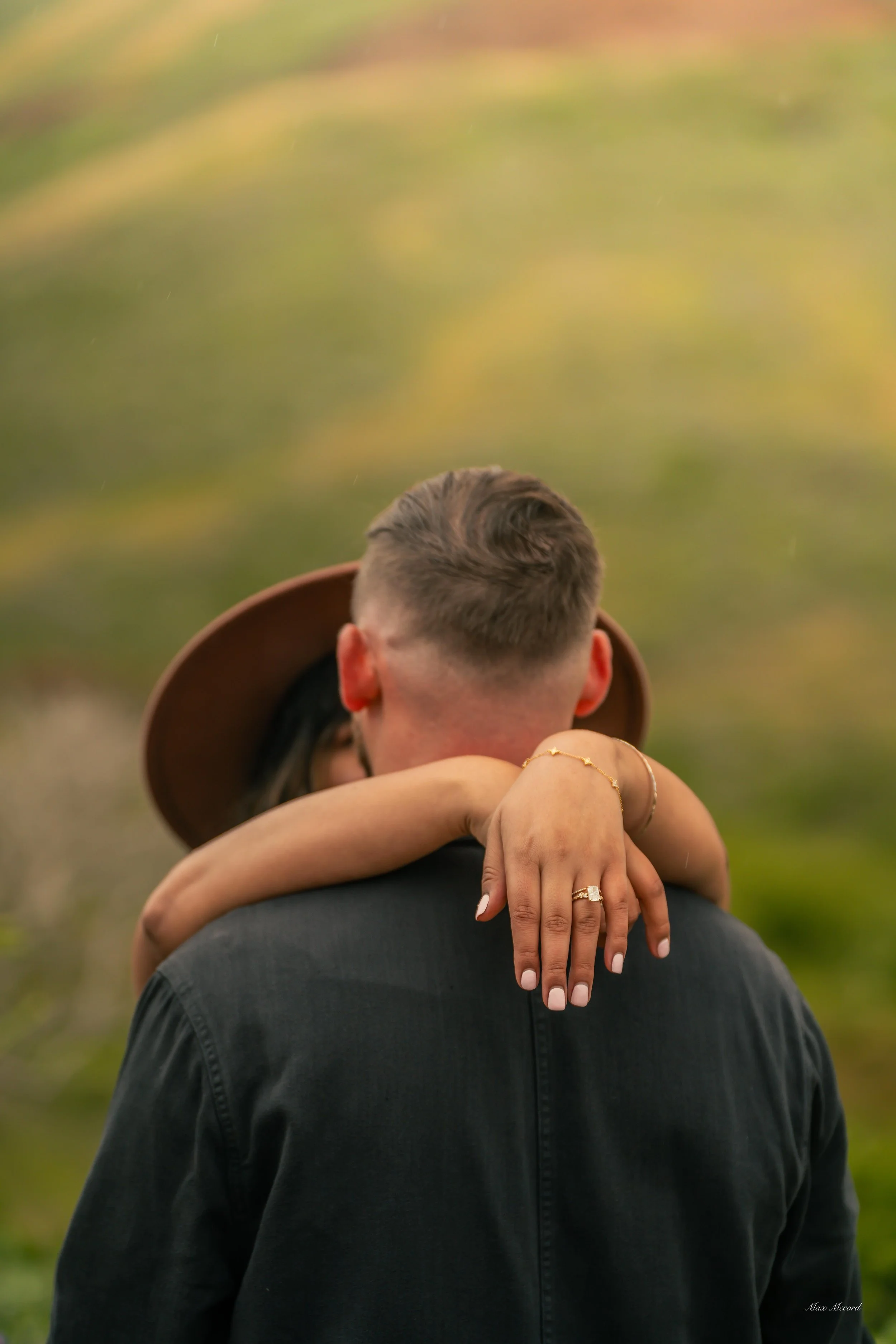 A man with short hair seen from behind, and a woman with her arms around his neck, hugging him. The woman's hands show her engagement ring and bracelet. The background is blurred greenery and a hillside.