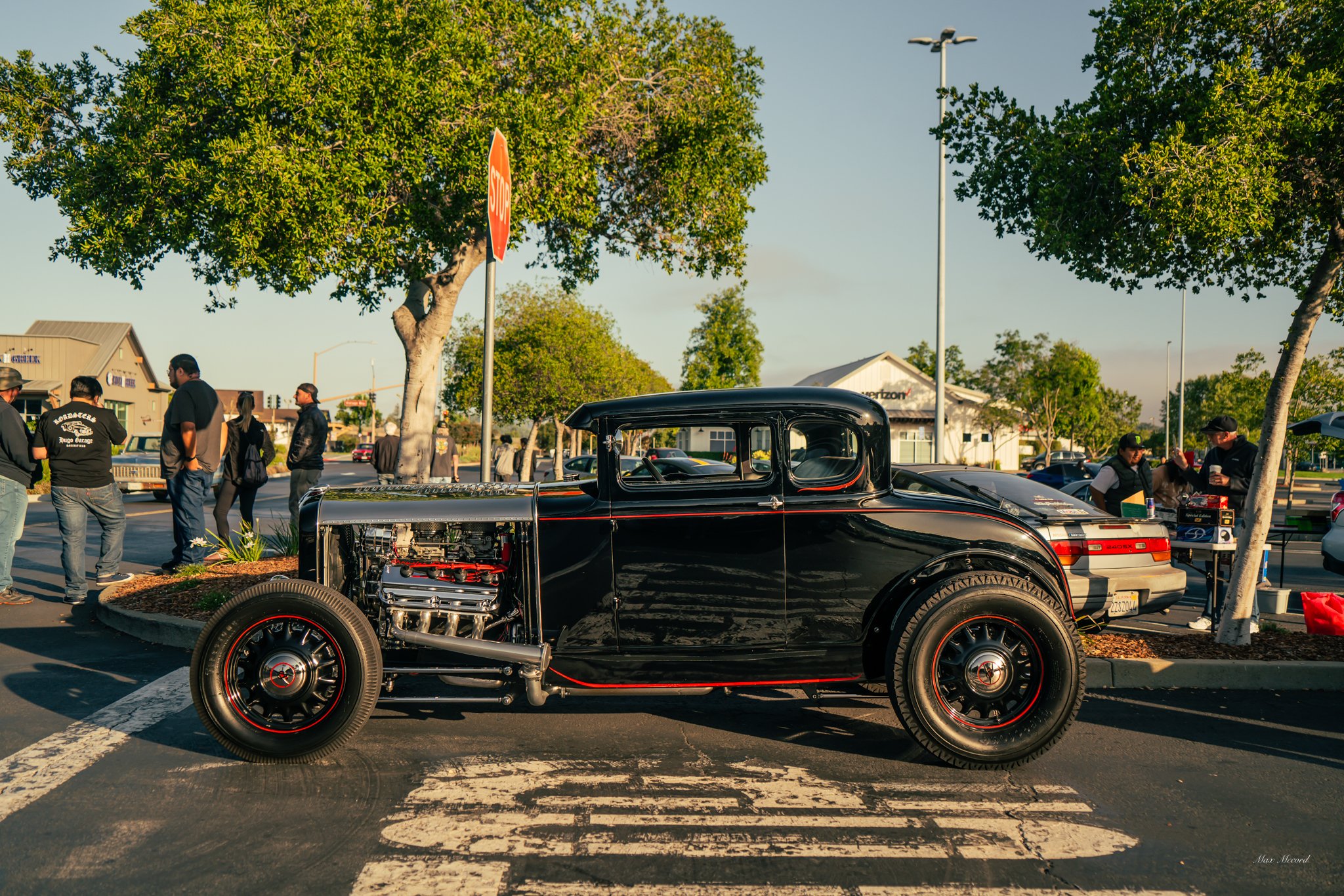 A vintage black hot rod car parked at an outdoor car show with people gathered around, trees, stop sign, and other cars in the background.