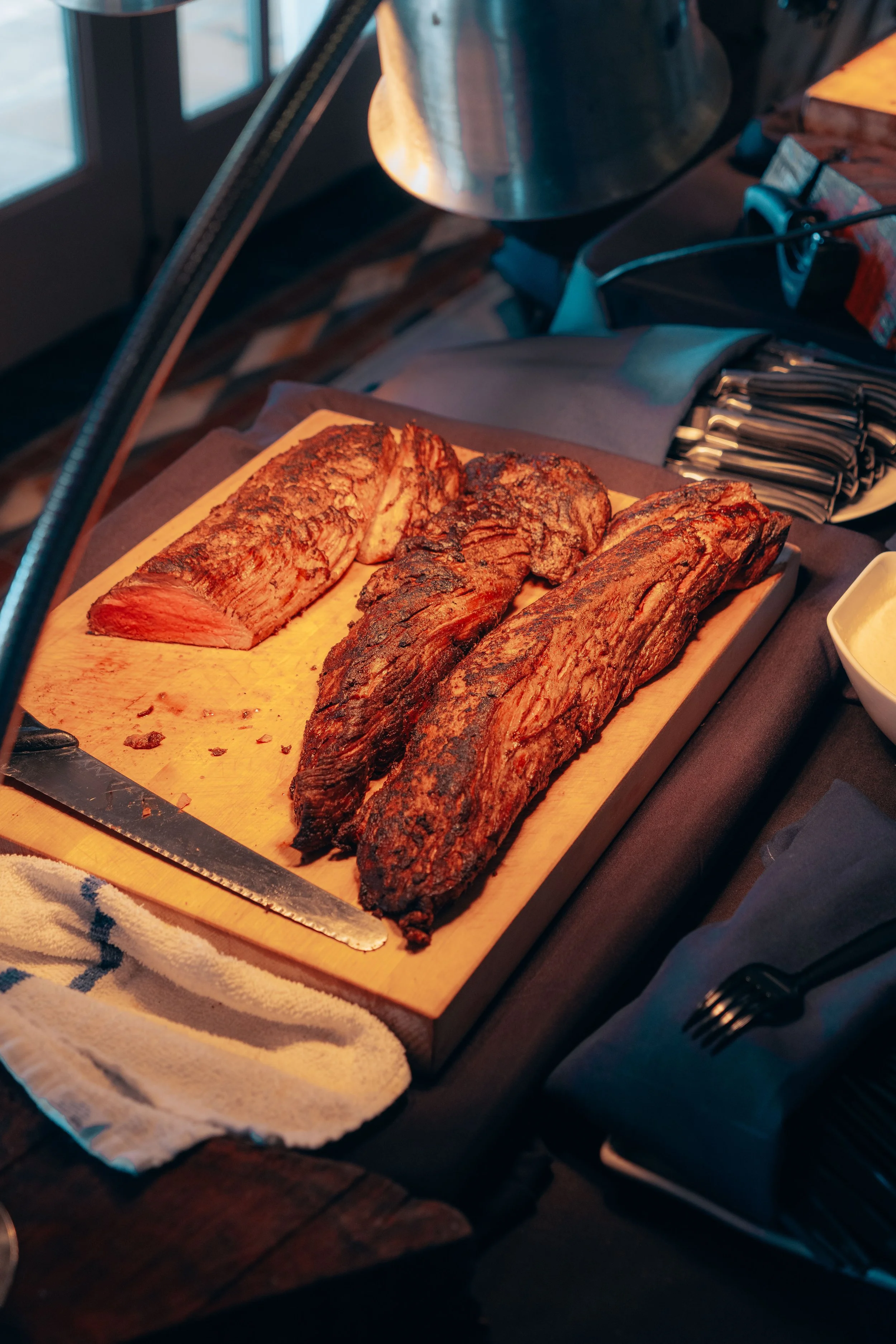 Smoked brisket meat on a wooden cutting board under a heat lamp, with a carving knife and a towel nearby.