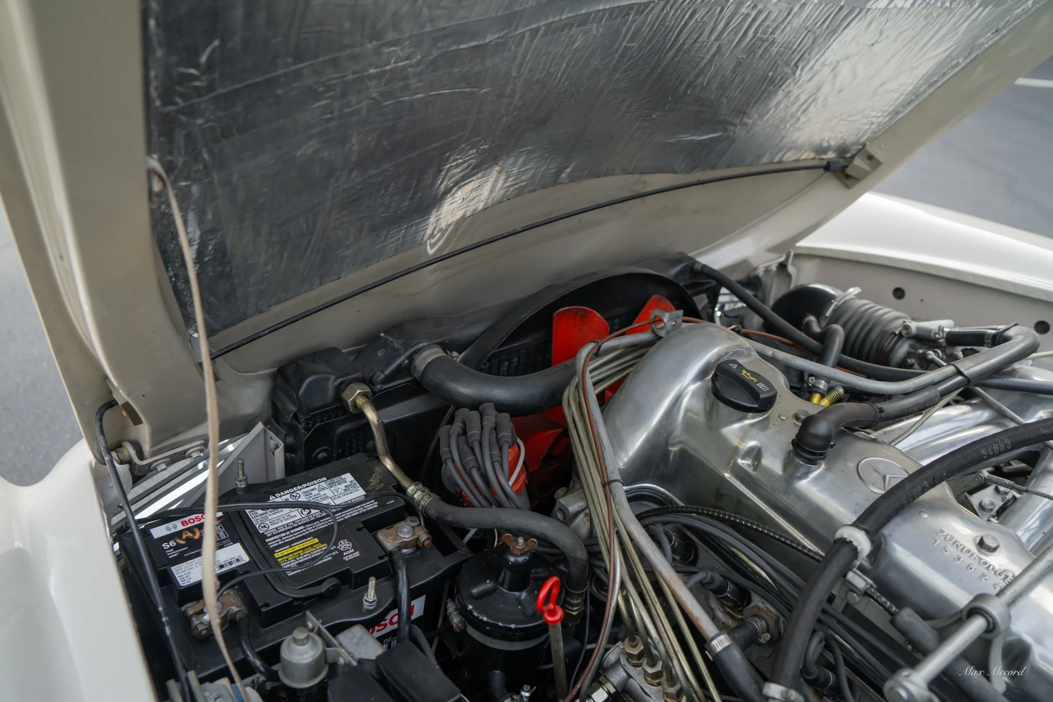 Close-up view of a vintage car engine with various hoses, wires, and metal components under the open hood.
