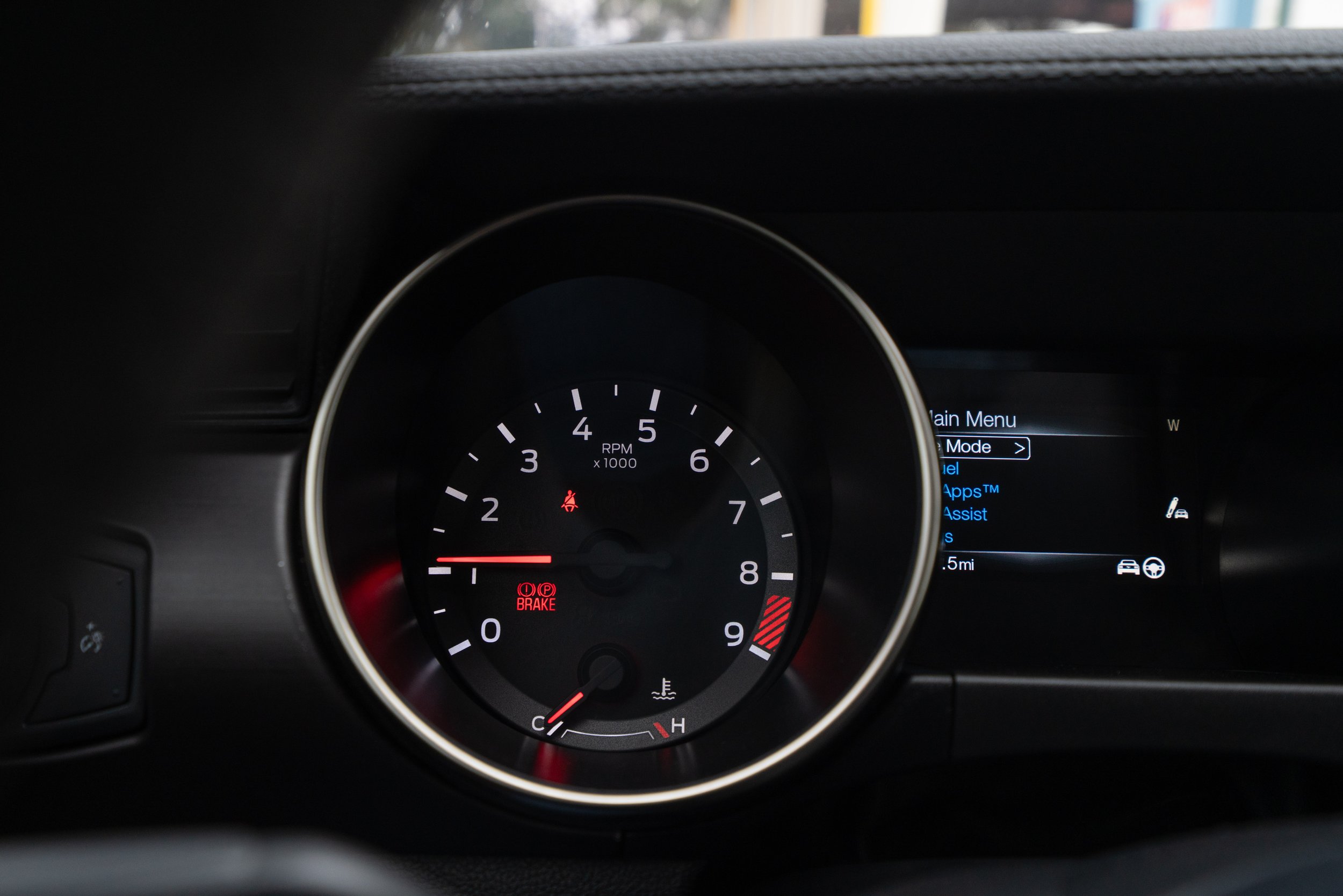 Close-up of a car's dashboard showing the tachometer with the needle pointing slightly below 1, the temperature gauge at cold, the brake warning light, and the seatbelt warning light. To the right, a digital screen displays menu options including mai