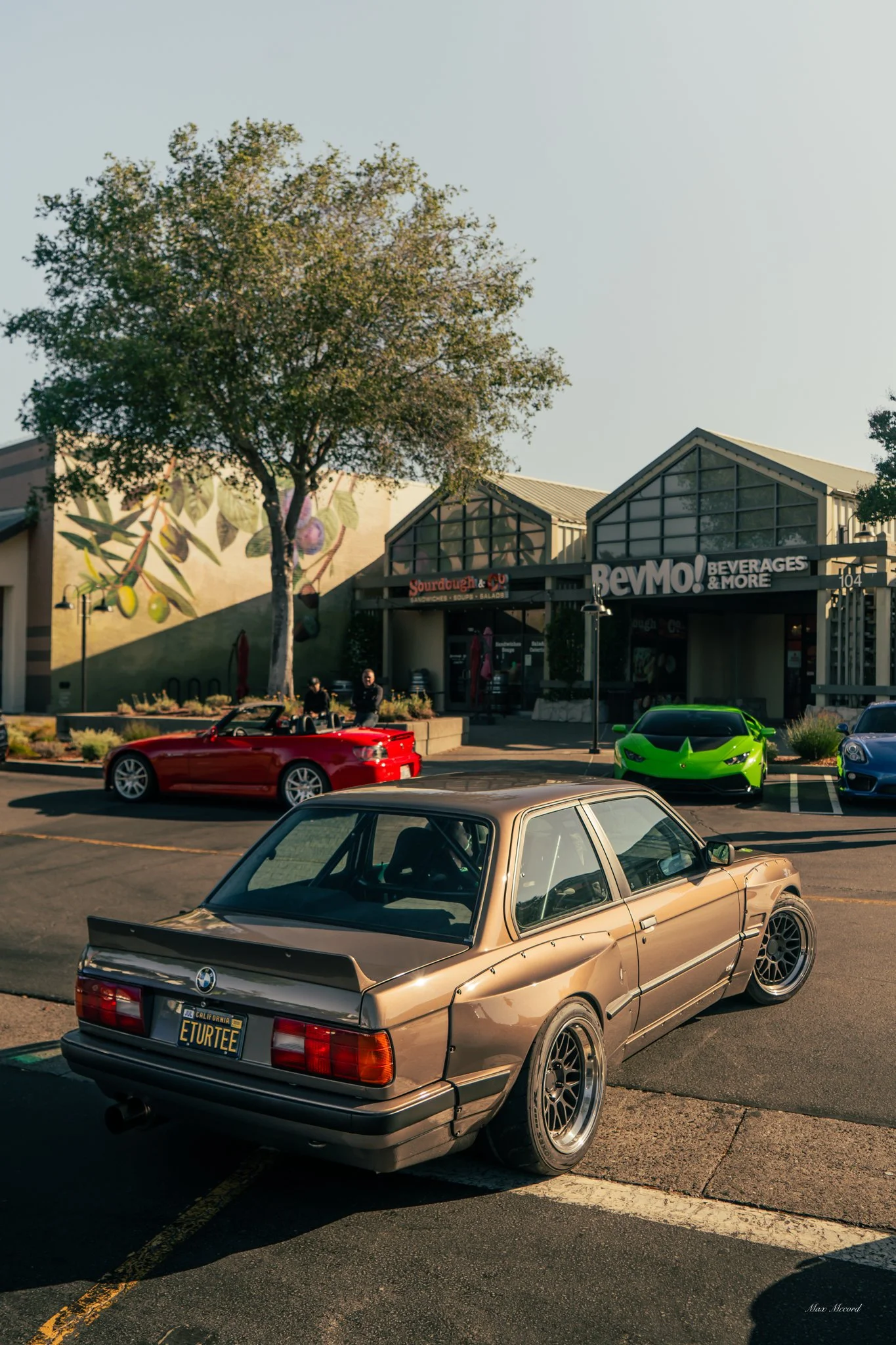 A vintage BMW E30 parked in a shopping center lot with other modern cars, a tree, and storefronts including a beverage and sourdough restaurant.