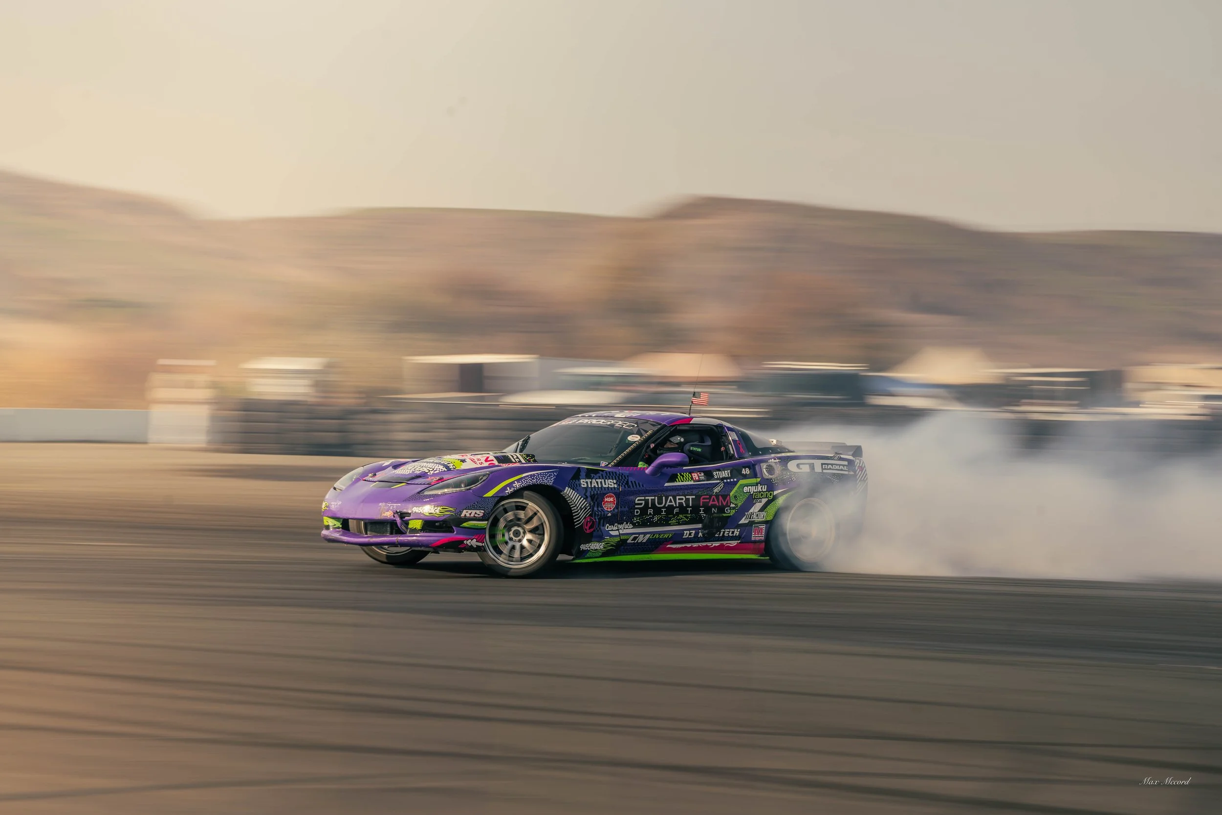 A purple and black race car drifting on a racetrack, creating smoke with a desert landscape in the background.