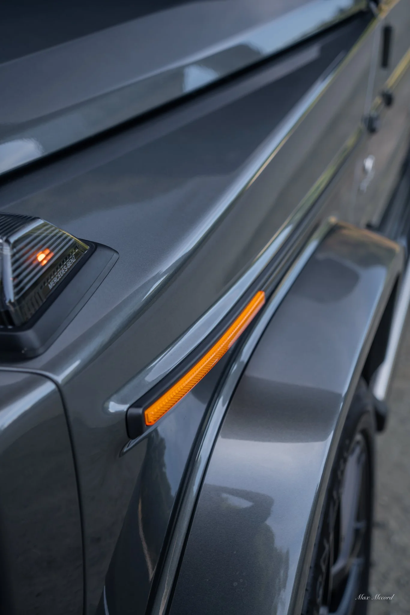 Close-up of the side of a dark gray luxury car, showing the hood, side mirror, and a part of the door with an orange side marker light.