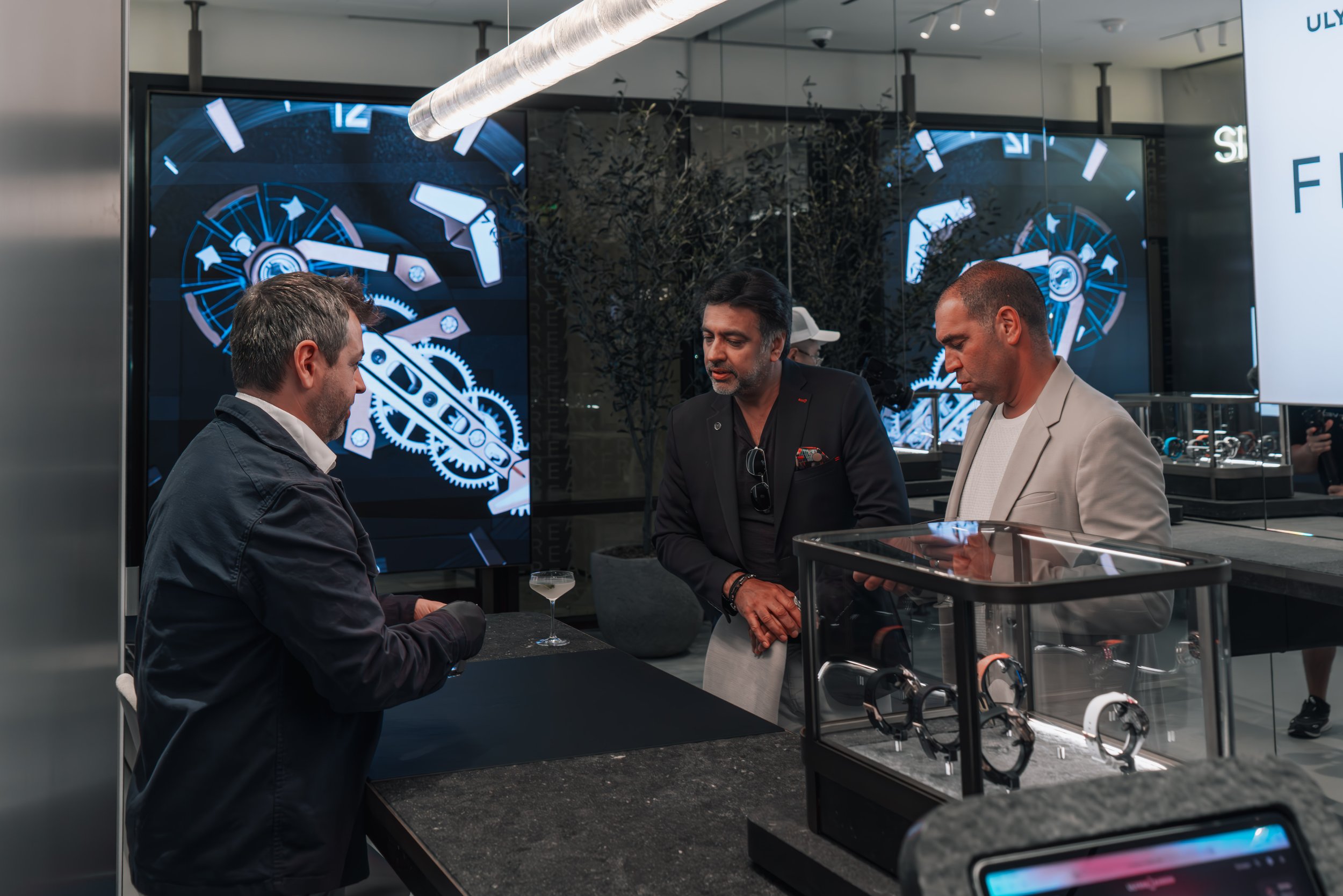 Three men standing at a counter in a watch store, looking at watches and a small glass of drink on the counter. There are large digital clocks on screens in the background and display cases with watches.