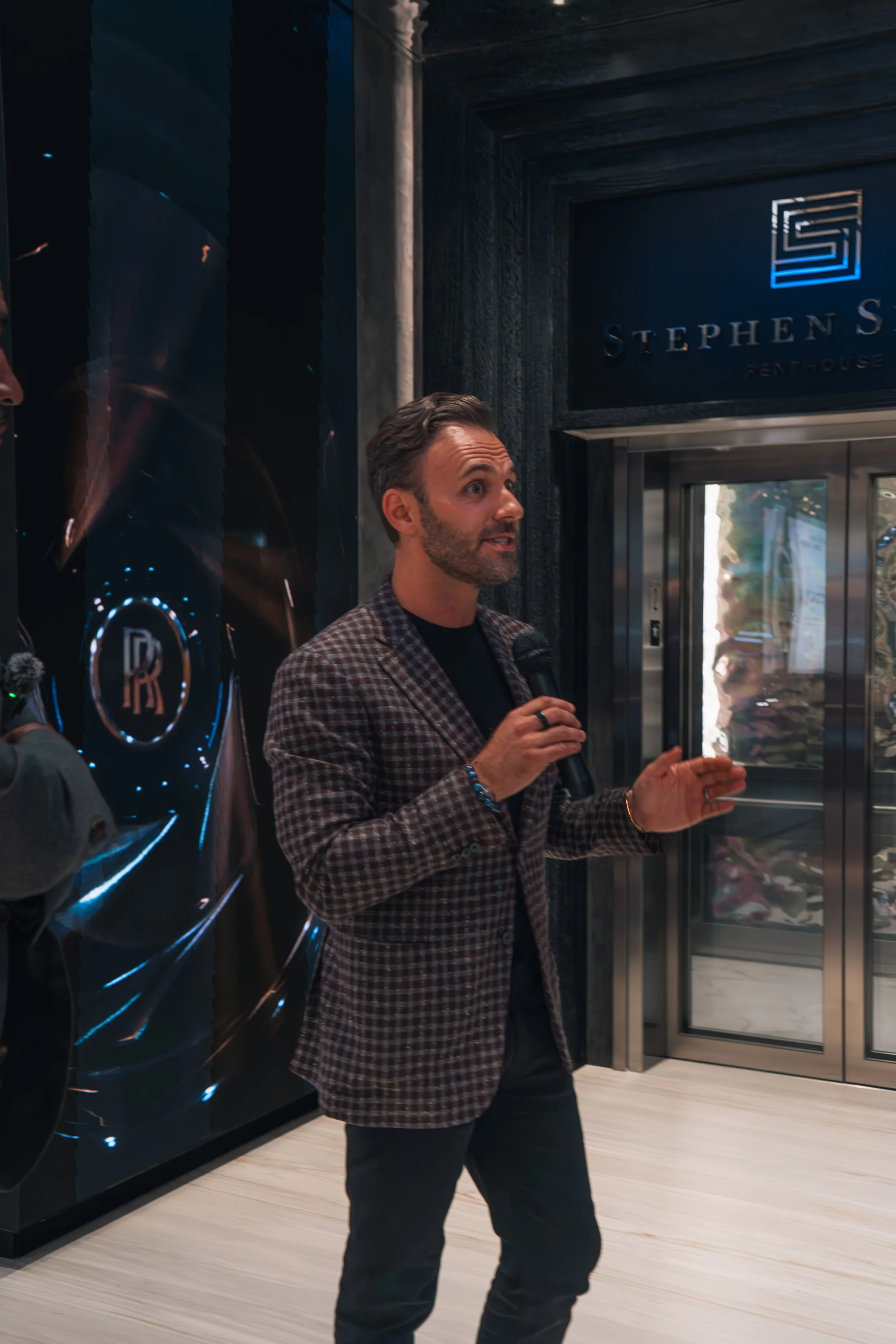 A man with dark hair and a beard, wearing a checkered blazer, holding a microphone, speaking in front of an elevator at Stephen S. Rosenthal Rentouise.