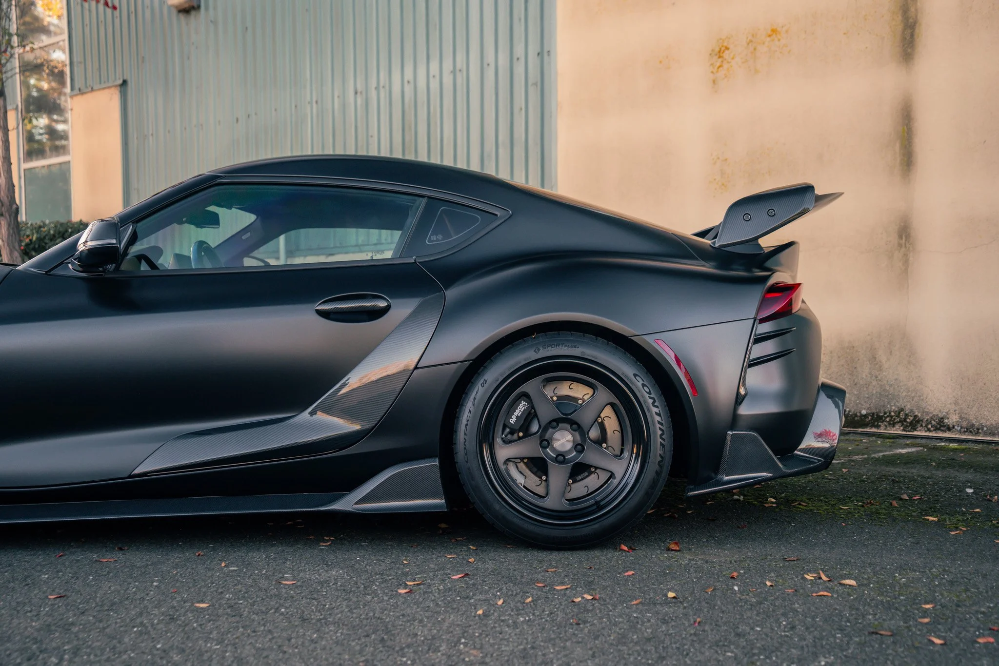 Black sports car with aerodynamic design, large rear wing, and racing tires parked on asphalt with fallen leaves near a textured wall.