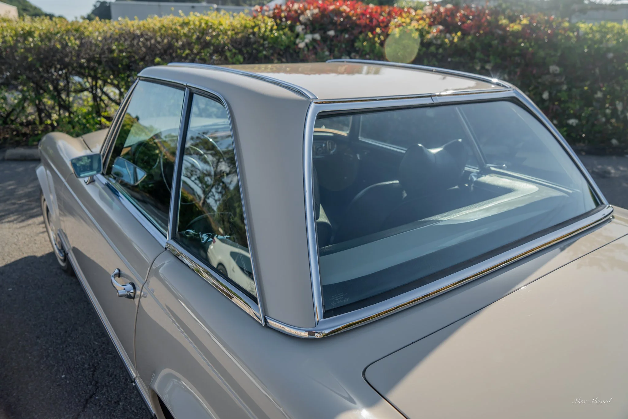 A vintage white car parked on the street, with a view of the windshield, side mirror, and part of the interior visible through the windows. There are bushes and flowers in the background.