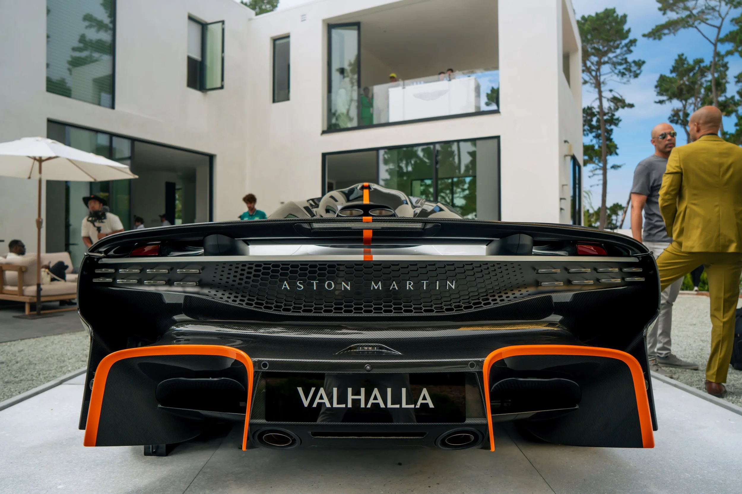 Rear view of a black Aston Martin Valhalla sports car with orange accents, parked outdoors near a modern white building, with people in the background